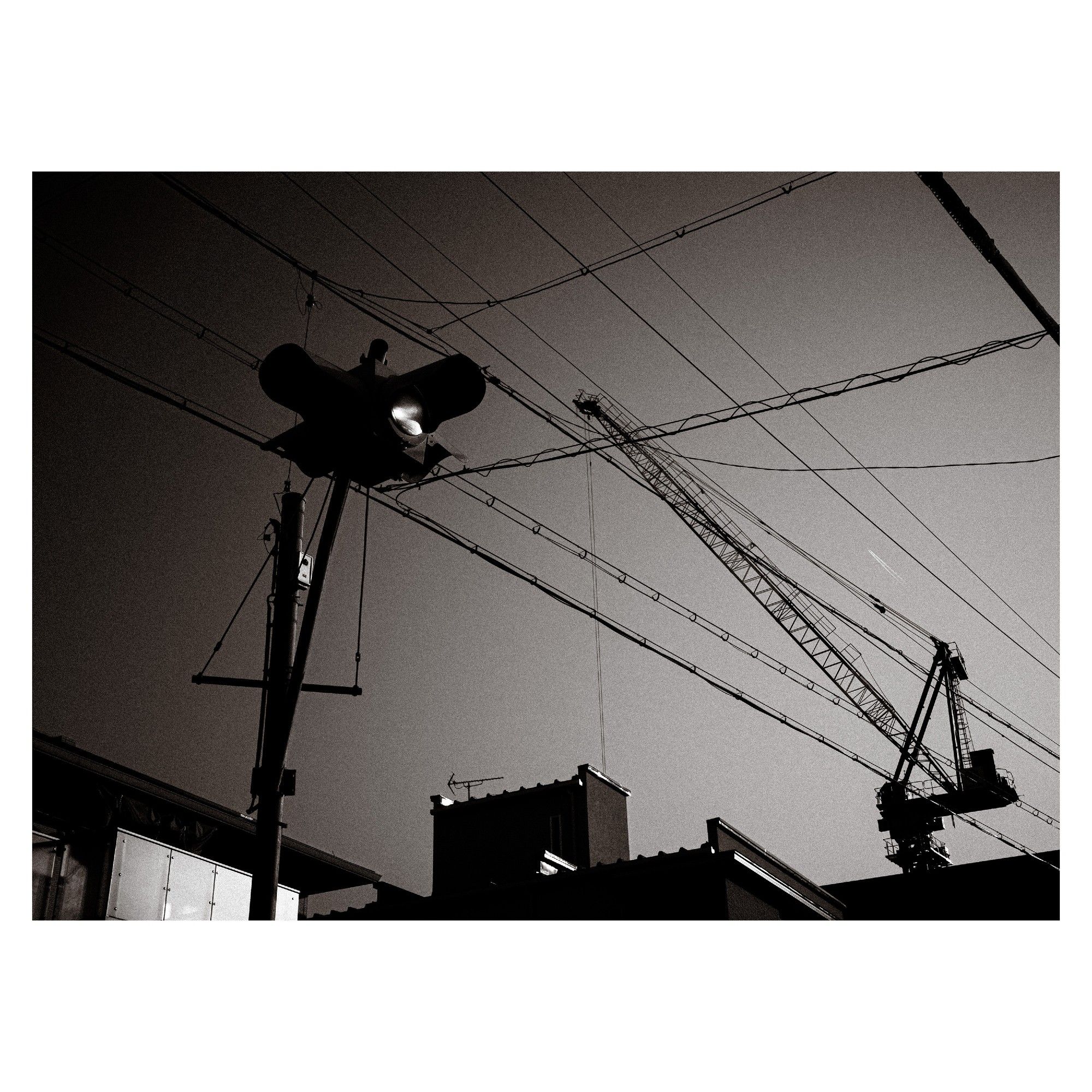 The black-and-white photo captures a traffic light in the foreground, slightly tilted and hanging from a pole. Above, a crisscross of power lines stretches across the sky, and in the background, two large construction cranes rise over rooftops, silhouetted against the evening sky. The image contrasts the static nature of the traffic light with the ongoing progress of construction, all framed by a tangle of urban infrastructure.