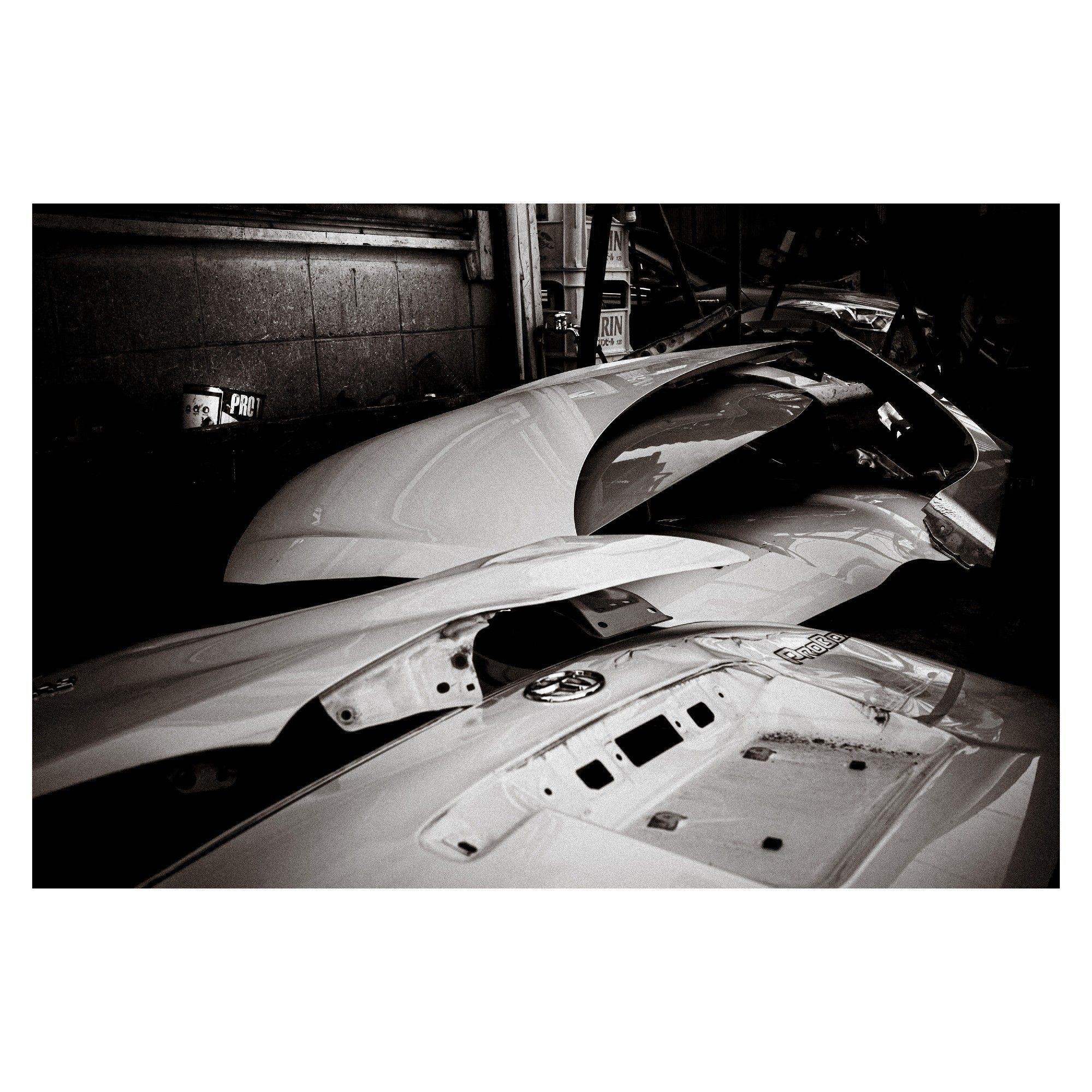 In this black and white photograph, various car body parts are arrayed with a sense of both disarray and artistry. Hoods, doors, and other automotive panels are stacked and leaned against each other, each carrying the potential to complete a vehicle. The scattered shadows and highlights across the metal surfaces suggest the warm ambiance of a spring day in a garage. There's a feeling of dormant energy, as if the parts are in a peaceful rest before coming together to form something whole and mobile once again.