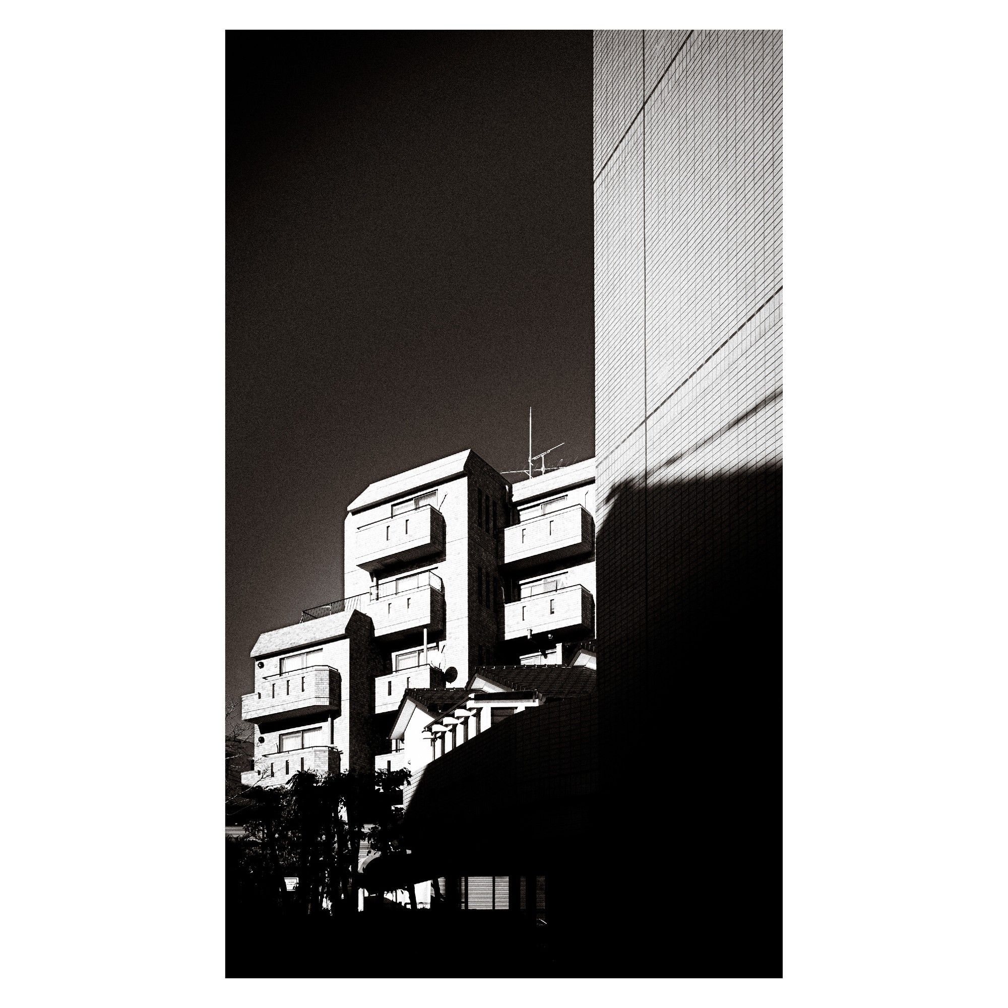 The black-and-white photo shows a tall, modern apartment building bathed in sharp sunlight. The structure has distinct geometric shapes, with deep balconies and sharp angles. A large shadow from another building falls across part of the frame, adding a stark contrast between light and darkness. The scene emphasizes the dramatic lines and architectural details of the building, giving it a powerful, almost looming presence against the dark sky.