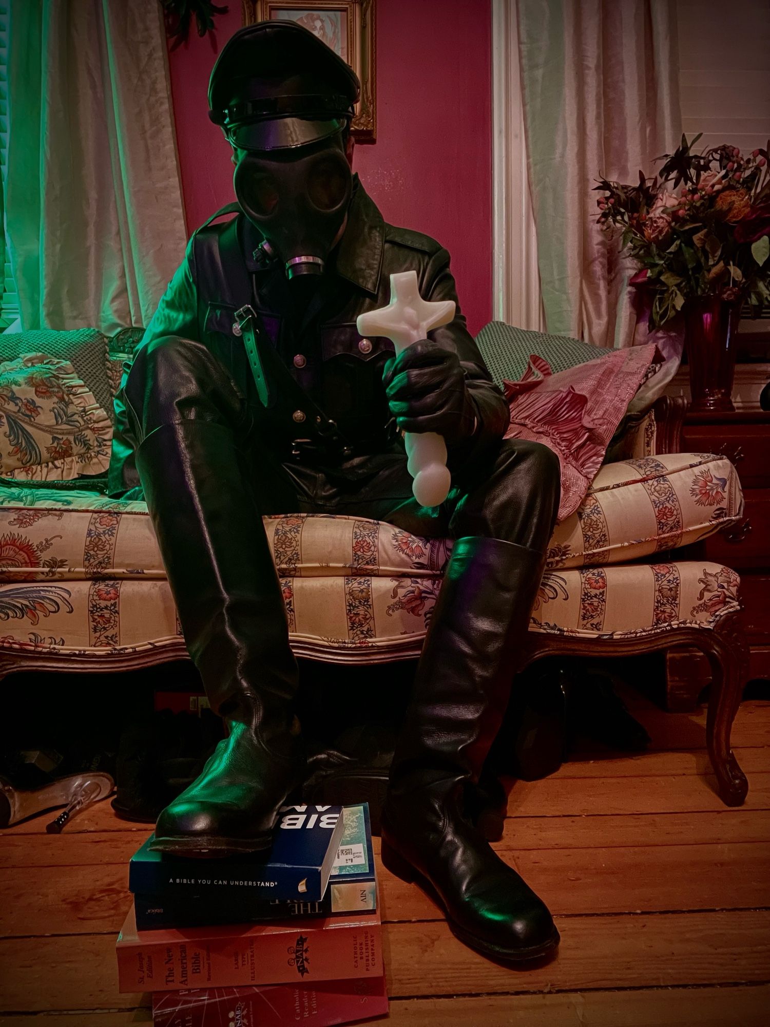 Image of a man in a black leather uniform, boots, gloves and cap, wearing a gas mask. He is sitting on an antique love seat next to a bouquet of flowers. He has one foot resting on a pile of Bibles and is holding a dildo shaped like a crucifix