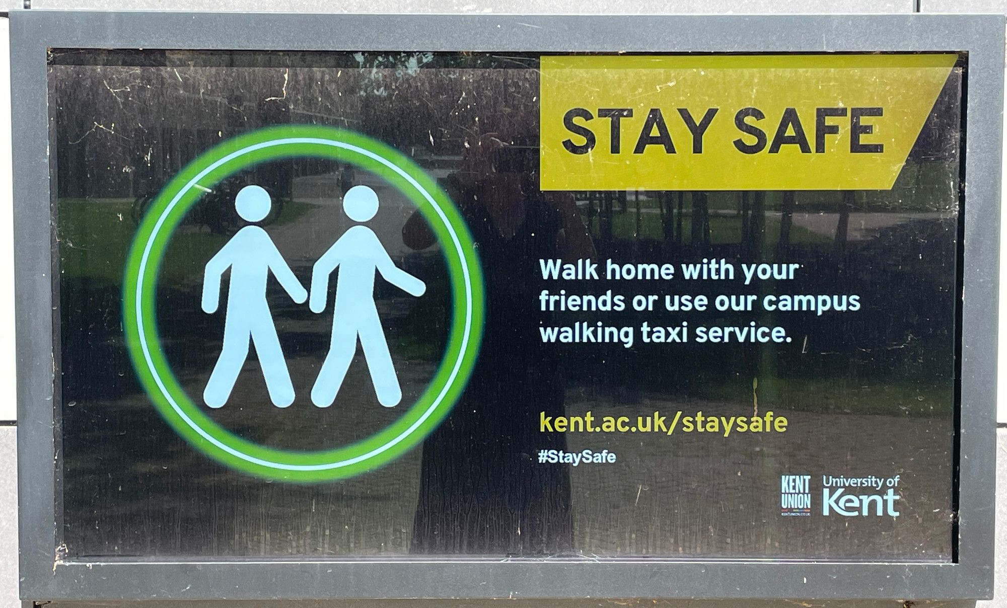 Foto eines Außenbildschirms vor dem Sicherheitsbüro der University of Kent (Querformat). Die Aufschrift lautet: Stay safe. Walk home with your friends or use our campus walking taxi service. kent.ac.uk/staysafe, #StaySafe. Auf der linken Seite des Bildschirms sind zwei stilisierte Personen in einem grünen Kreis abgebildet, die gemeinsam gehen.