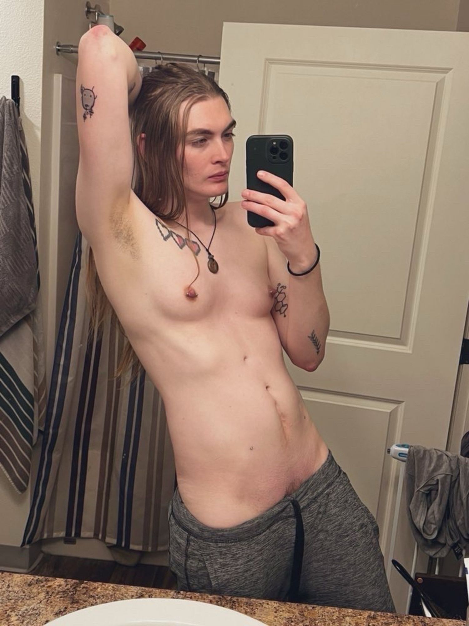 Zai shirtless with her long brown hair takes a mirror selfie in a bathroom, raising one arm and holding a phone in the other. She is wearing gray sweat pants and there are towels and toiletries in the background.