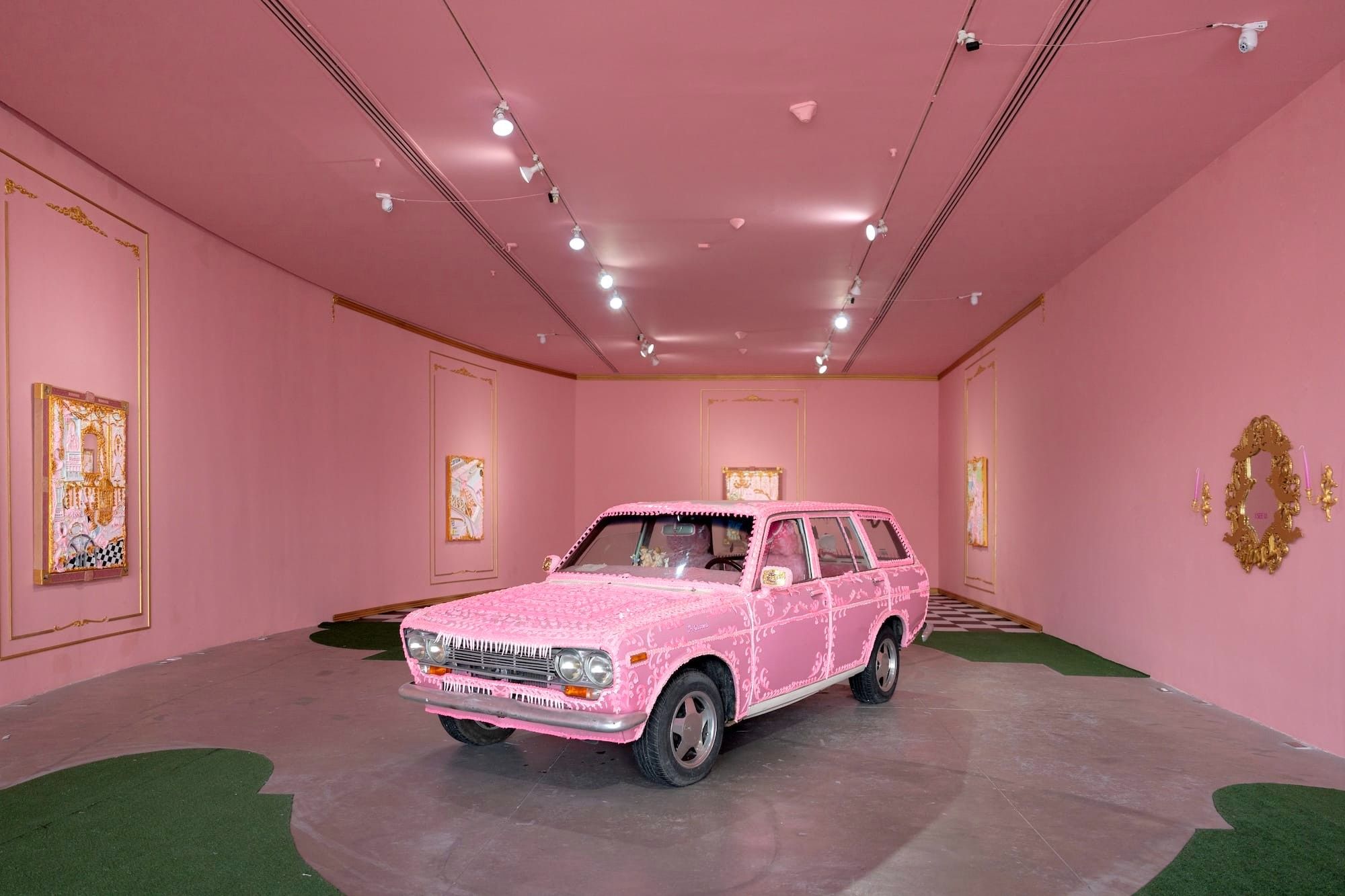 an installation view of an exhibition featuring pink walls, green turf on the floor, and a pink Datsun in the center