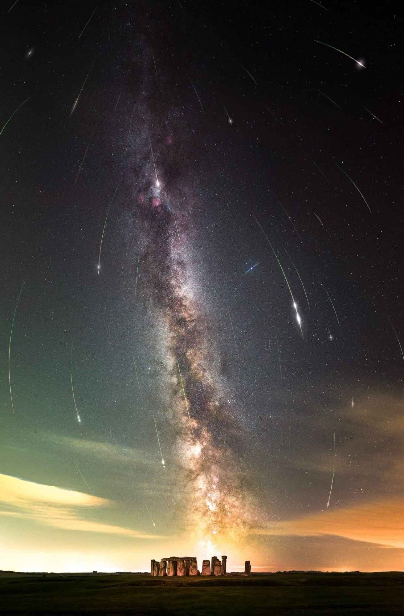 meteors streak down the sky to frame the milky way, while stonehenge sits at the base