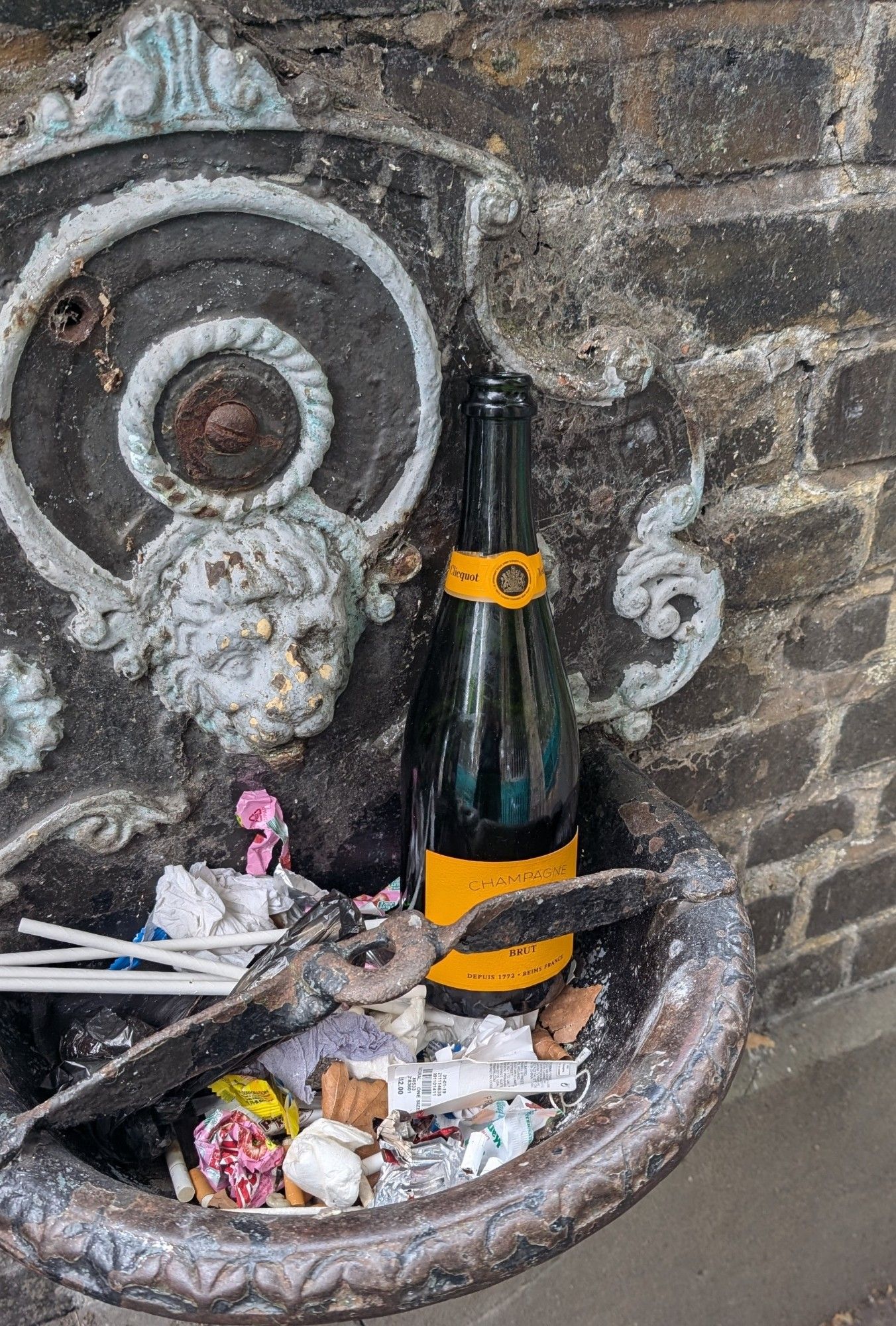 Victorian stone water fountain with lion's mouth into scallop edged bowl. Bowl filled with assorted small pieces of rubbish. And an empty bottle of Veuve Cliquot champagne.