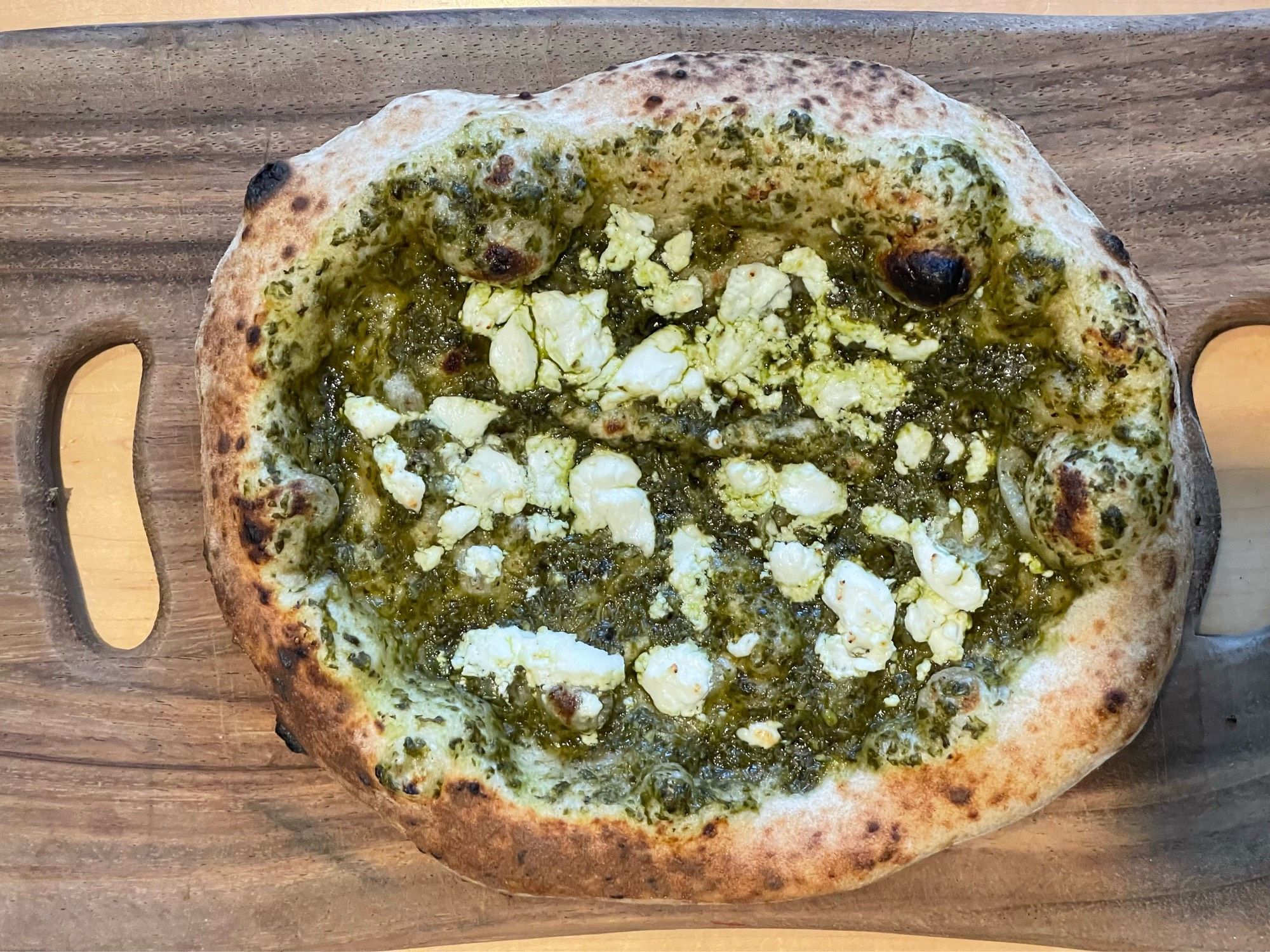 A 12-i inch pizza with green pesto sauce and white goat cheese, with a golden brown crust