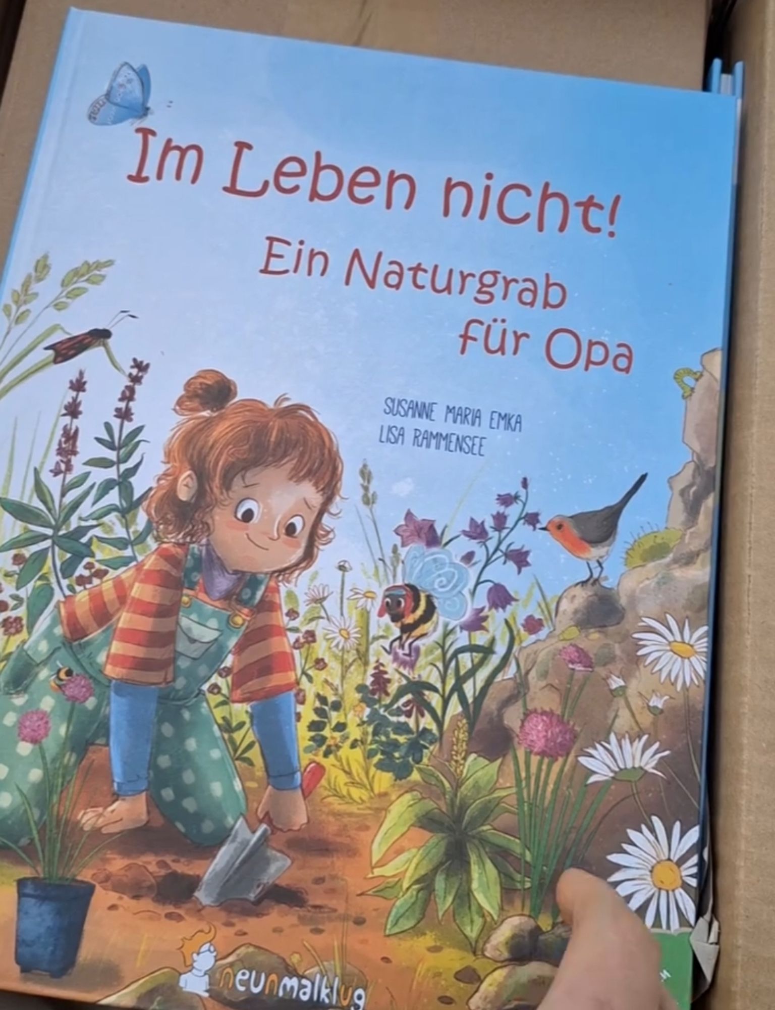 Foto vom Buch "Im Leben nicht! Ein Naturgrab für Opa" von Susanne Maria Emka und Lisa Rammensee
Auf dem Cover sieht man ein Mädchen mit bunter Kleidung inmitten von Wildblumen und Insekten und Vögel knien. Das Mädchen schaufelt gerade ein Pflanzloch für eine Wildblume, die im Topf neben ihr steht.
Rechts unten an der Buchecke sieht man meinen Zeigefinger.