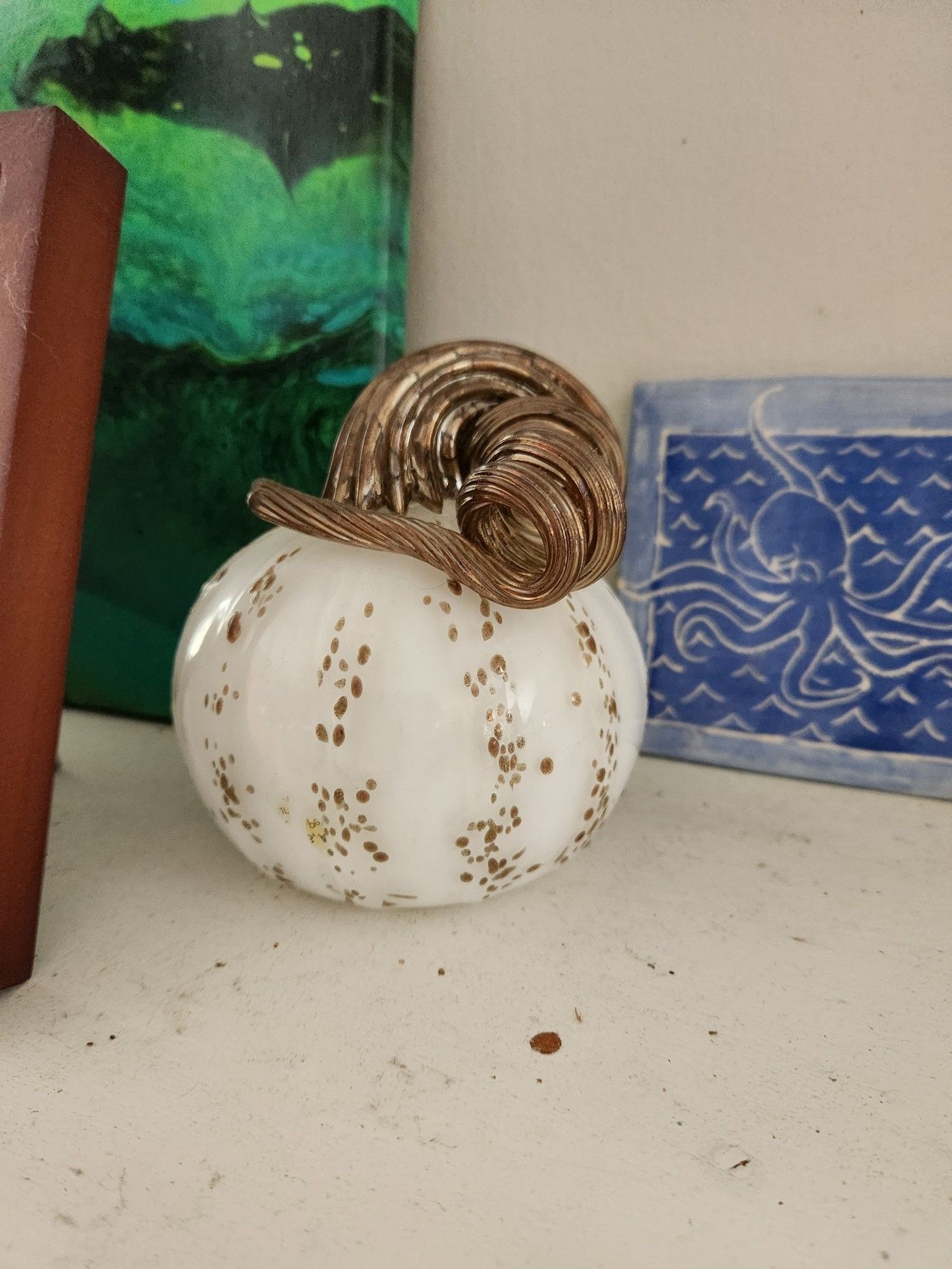 A white and gold blue  glass pumpkin on a scuffed white mantel; also visible are a green marbled canvas and a blue ceramic tile with an octopus on it