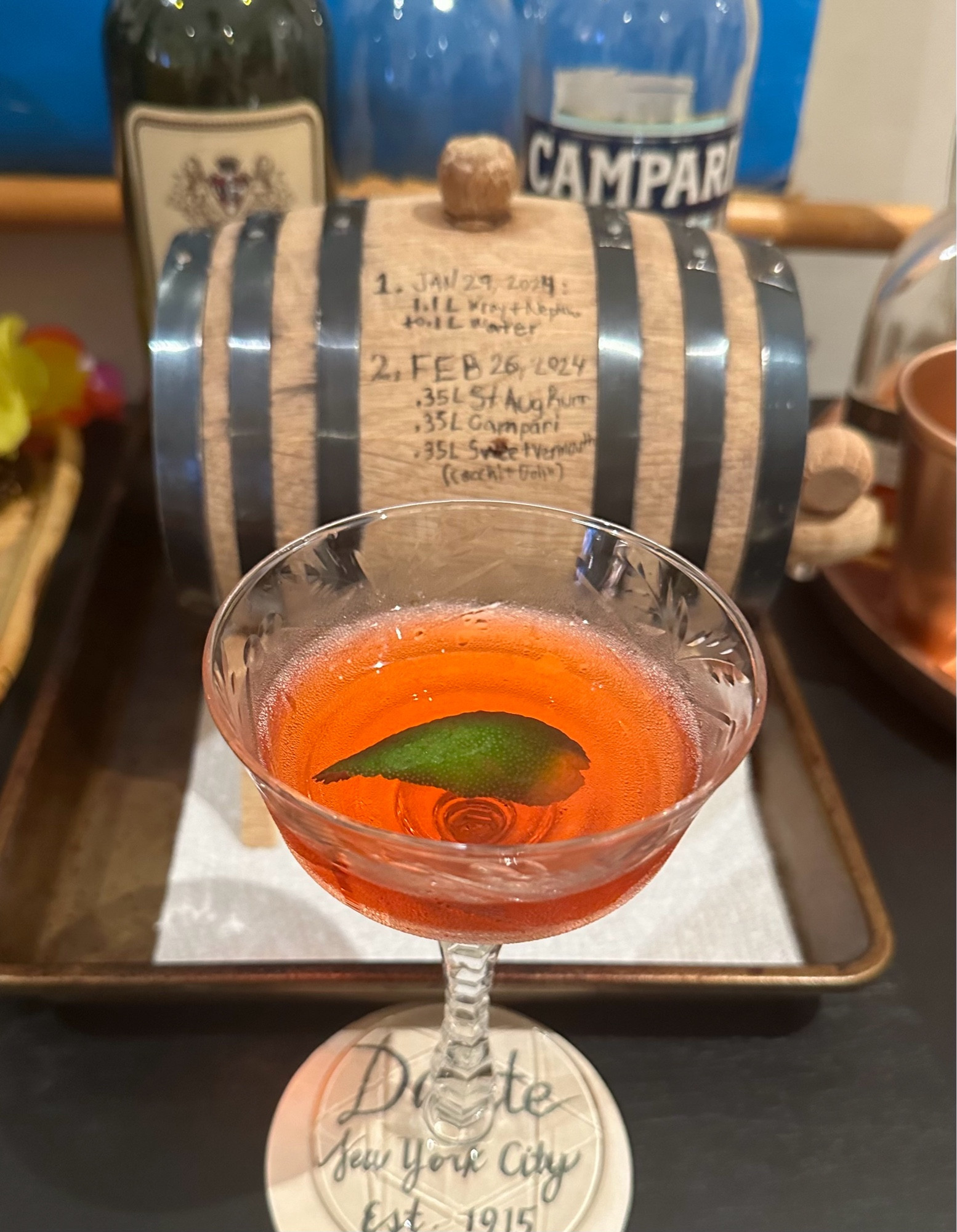 A rum negroni in a coupe glass, with a lime peel, in front of a mini barrel.