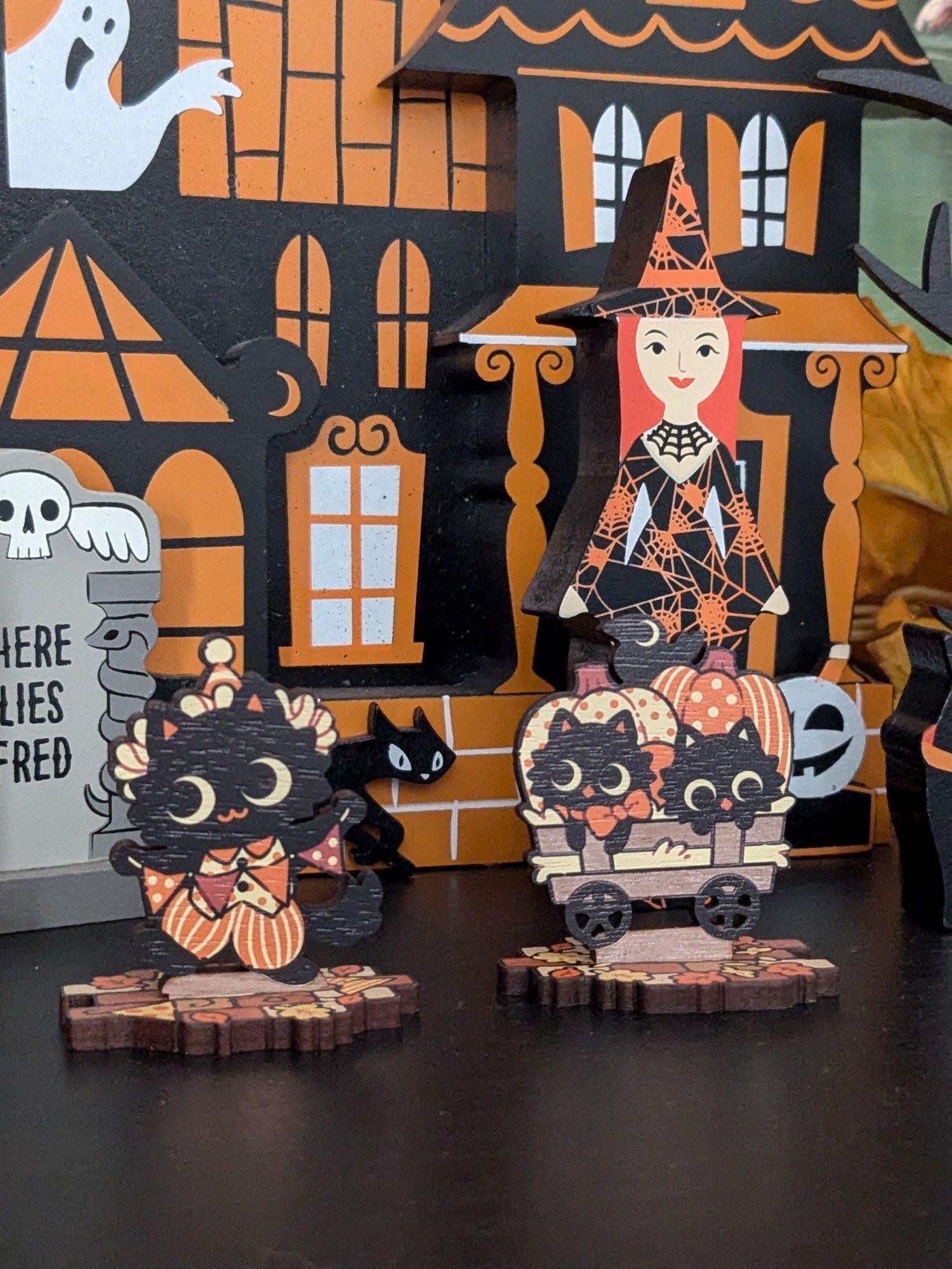 Photo of some wooden Halloween decorations sitting on a black mantle. There's a spooky house and witch in the back, with two cat-rhemed standees in the front - most recent purchases from Sugar Bunny Shop! The one on the left has a very clown-themed costume while the one on the right is a wagon with two small cats peaking out