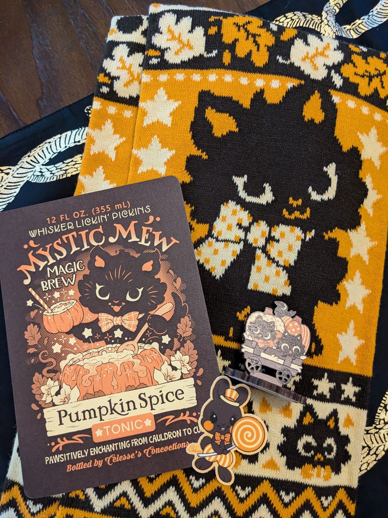 Photo of the goodies I ordered from Sugar Bunny Shop! There's an orange scarf in the back with a cute kitty on it, a print that is styled as an ad for mystic mews pumpkin spice tonic, again with a black kitty as the center piece, a black bunny holding an orange swirled lollipop, and the wagon standee with two tiny cats on it.