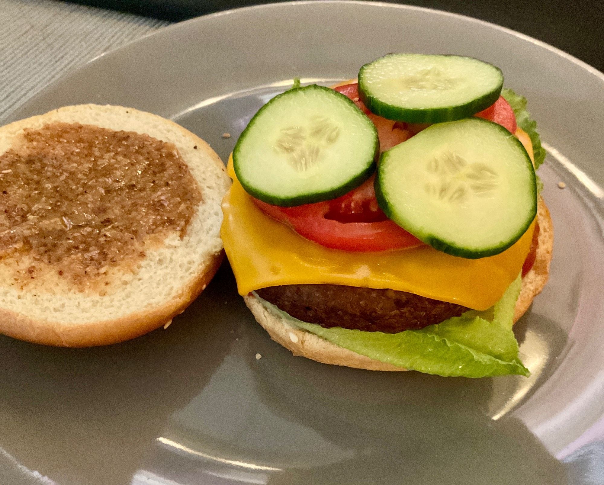 A vegan burger on a gray plate. Sweet mustard on the top bun. Cucumber slices, tomato, vegan cheddar, a soy-based patty, lettuce and ketchup.