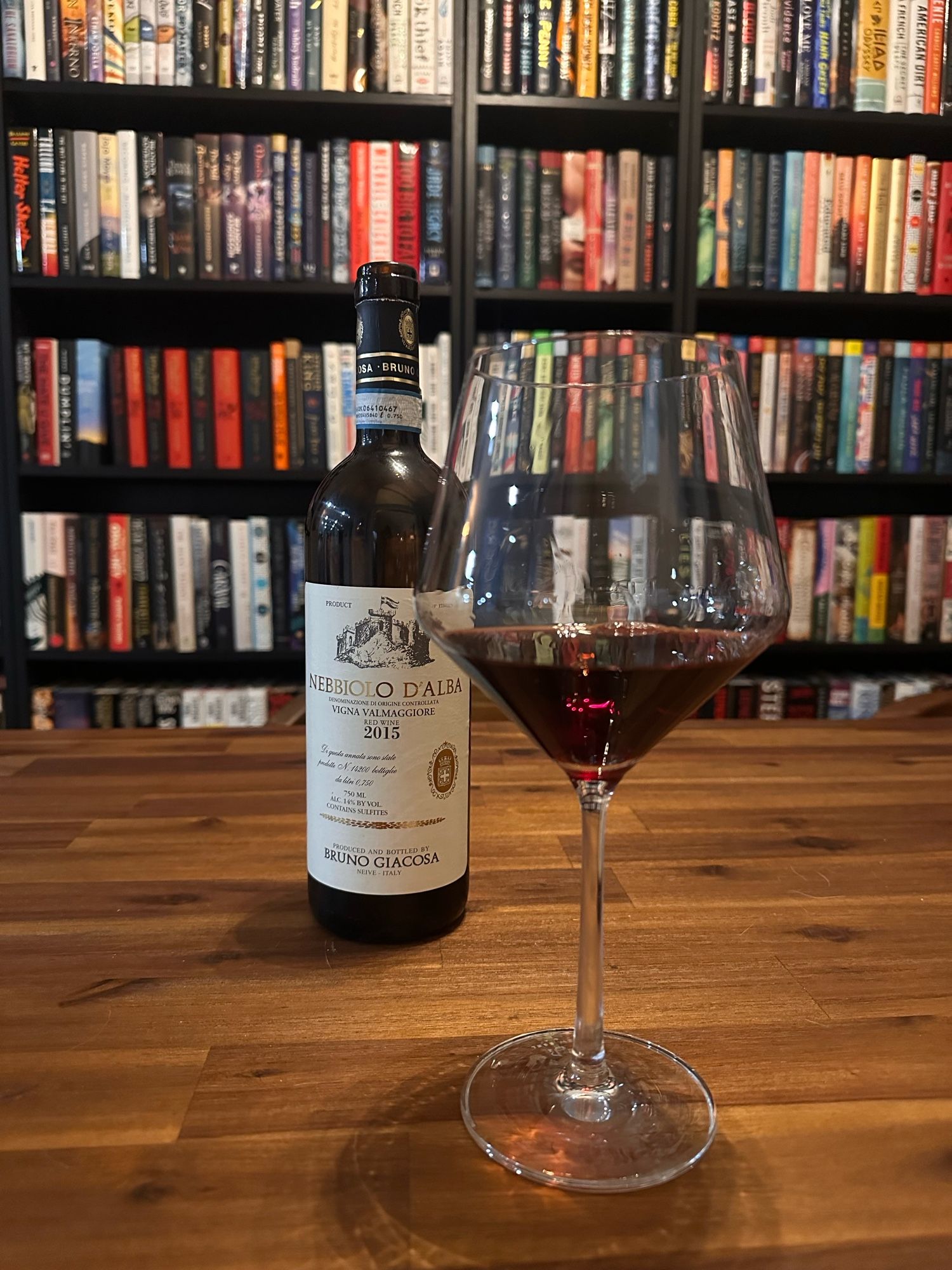 A glass of red wine next to a bottle of 2015 Nebbiolo d’alba in front of a book shelf