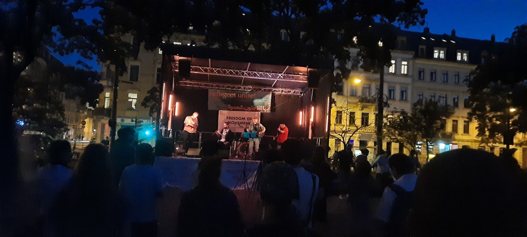 Eine Bühne, auf der Menschen mit Instrumenten stehen. Es gibt kurdische Musik. Vor der Bühne tanzen Leute in der Dämmerung.