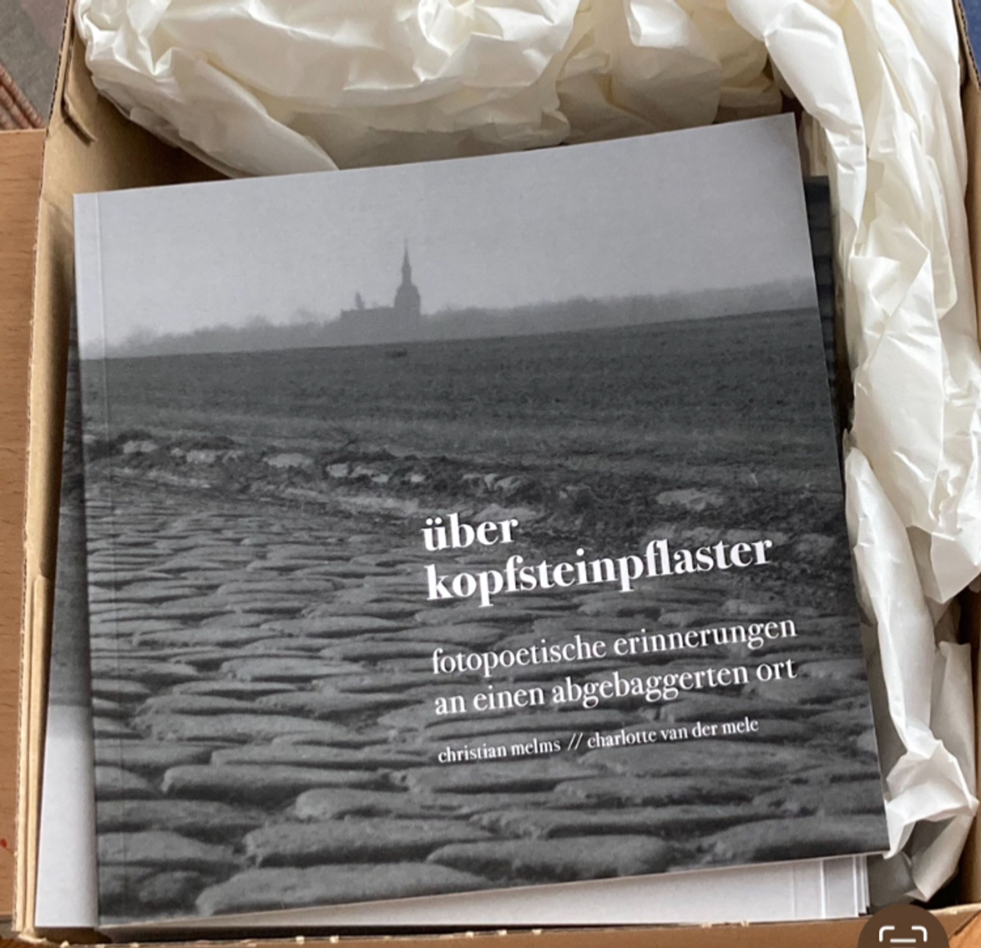 Die angelieferten Bücher noch in der Kiste mit Packpapier.
Das Cover ist schwarz/weiß. Eine Kopsteinpflasterstraße und in der Ferne eine Kirche und Bäume.