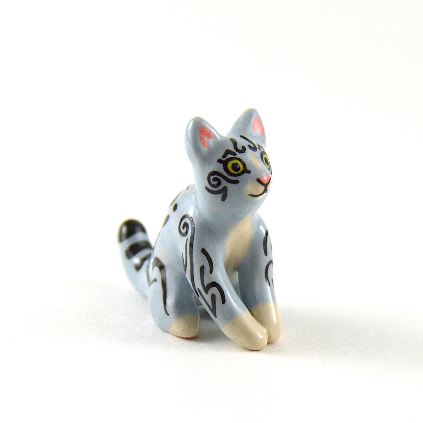 A picture of a small ceramic cat. The cat is sitting and is painted like a gray tabby with green eyes.