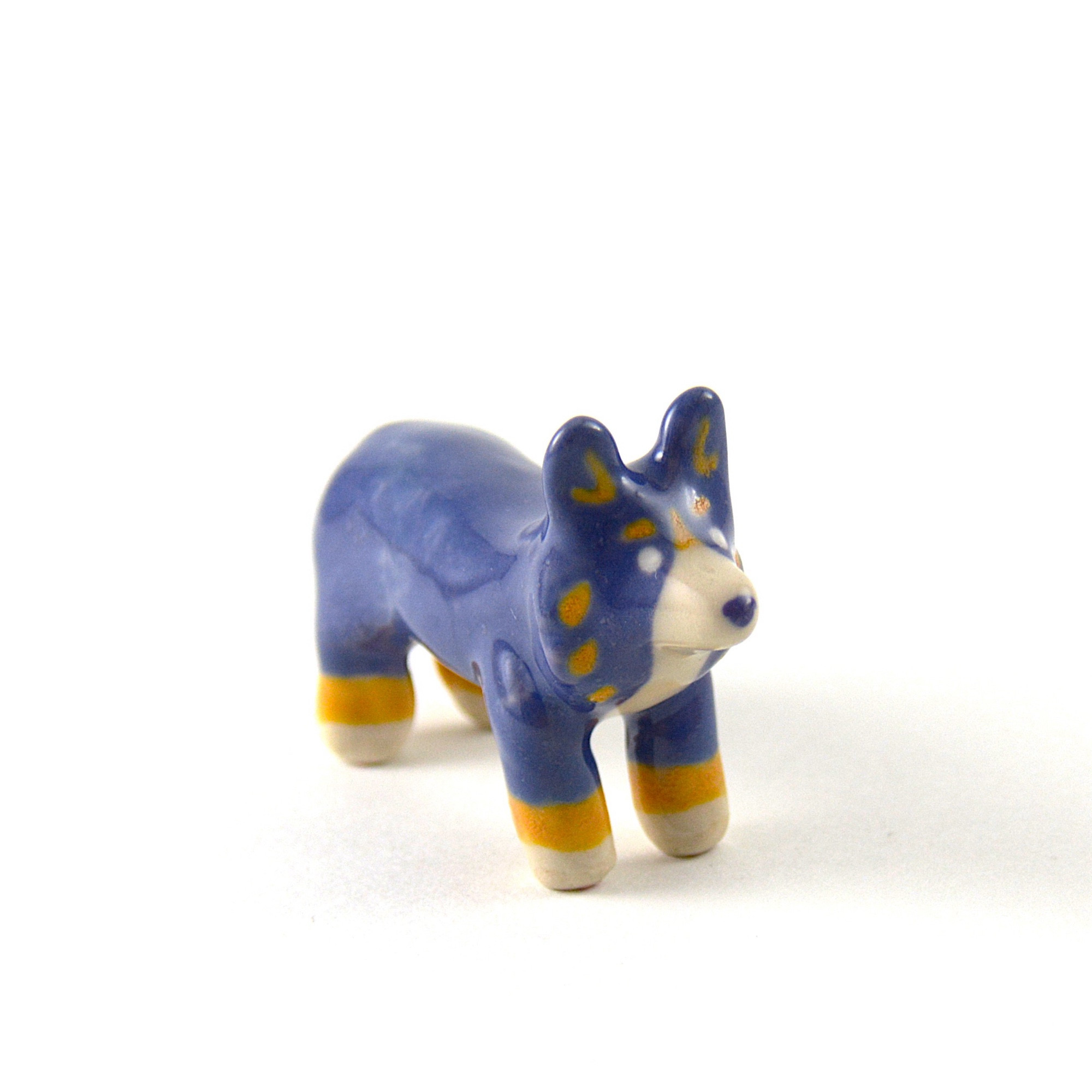 A little ceramic corgi sculpture on a white background. This corgi is painted purple with orange and white accents.