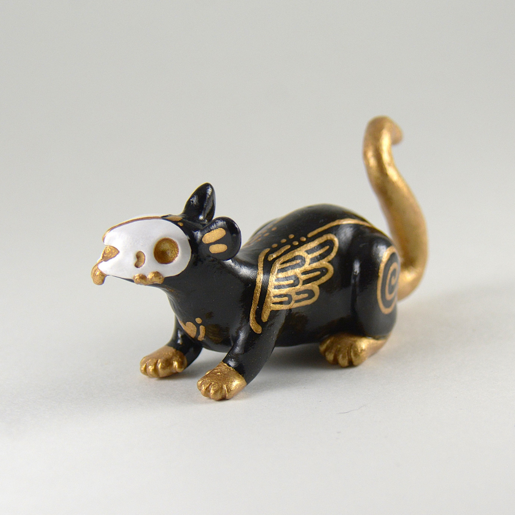 A little rat sculpture on a white background. The rat is painted black with gold details and it has a skull for a face.