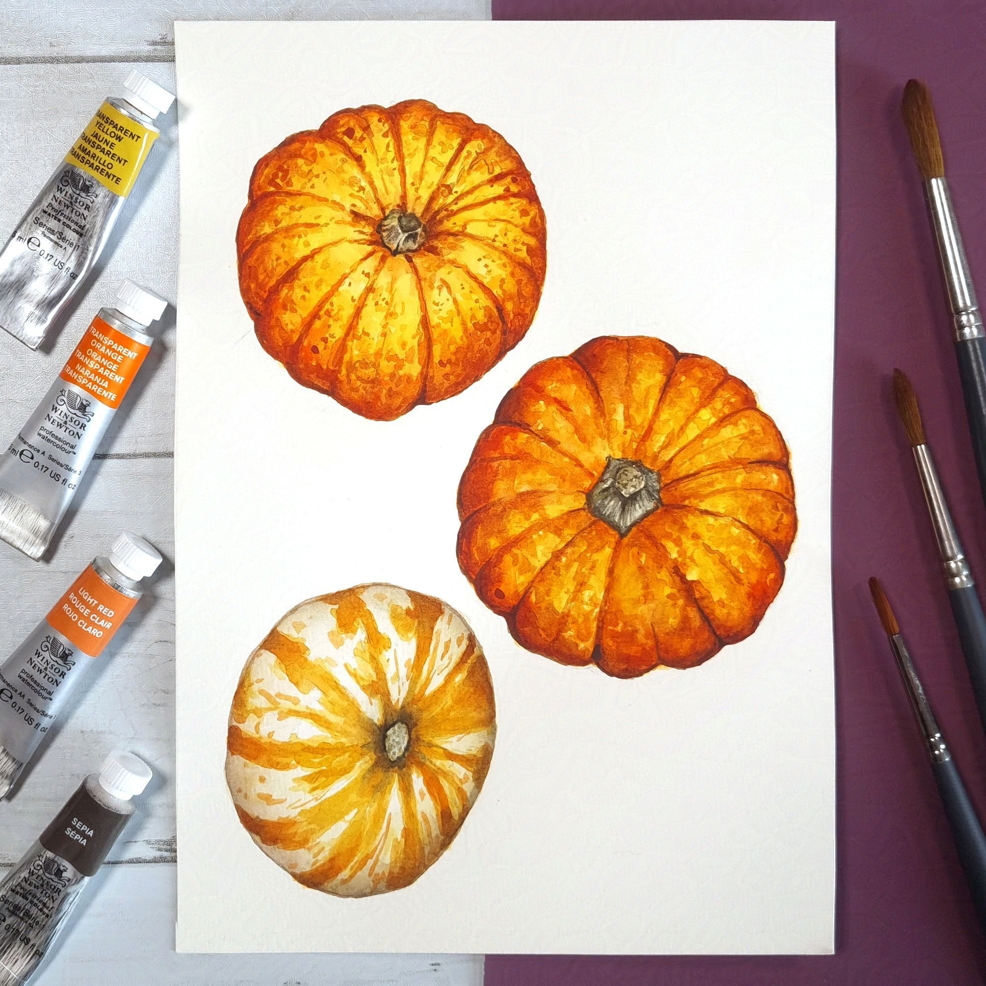 three orange pumpkin paintings in watercolour, paint tubes on right and brushes to the left