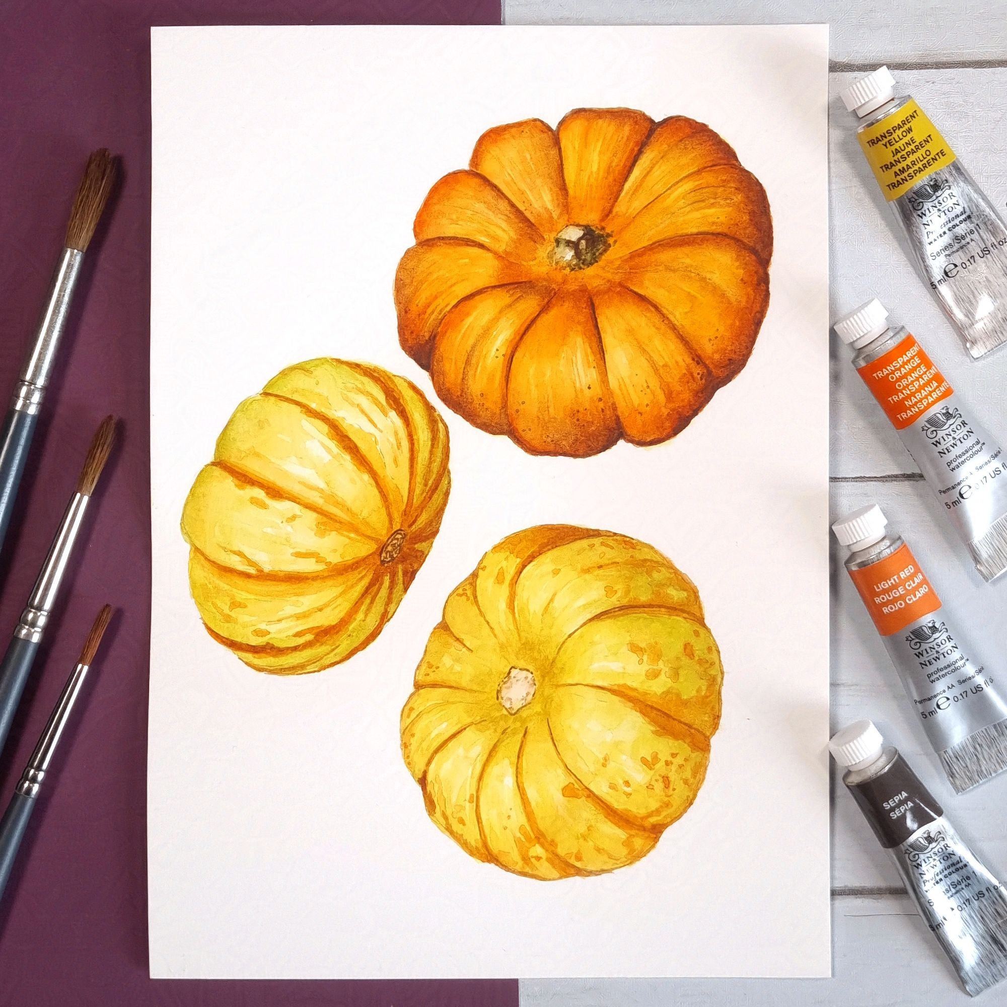 3 orange pumpkin paintings, paint tubes on right and brushes on the left