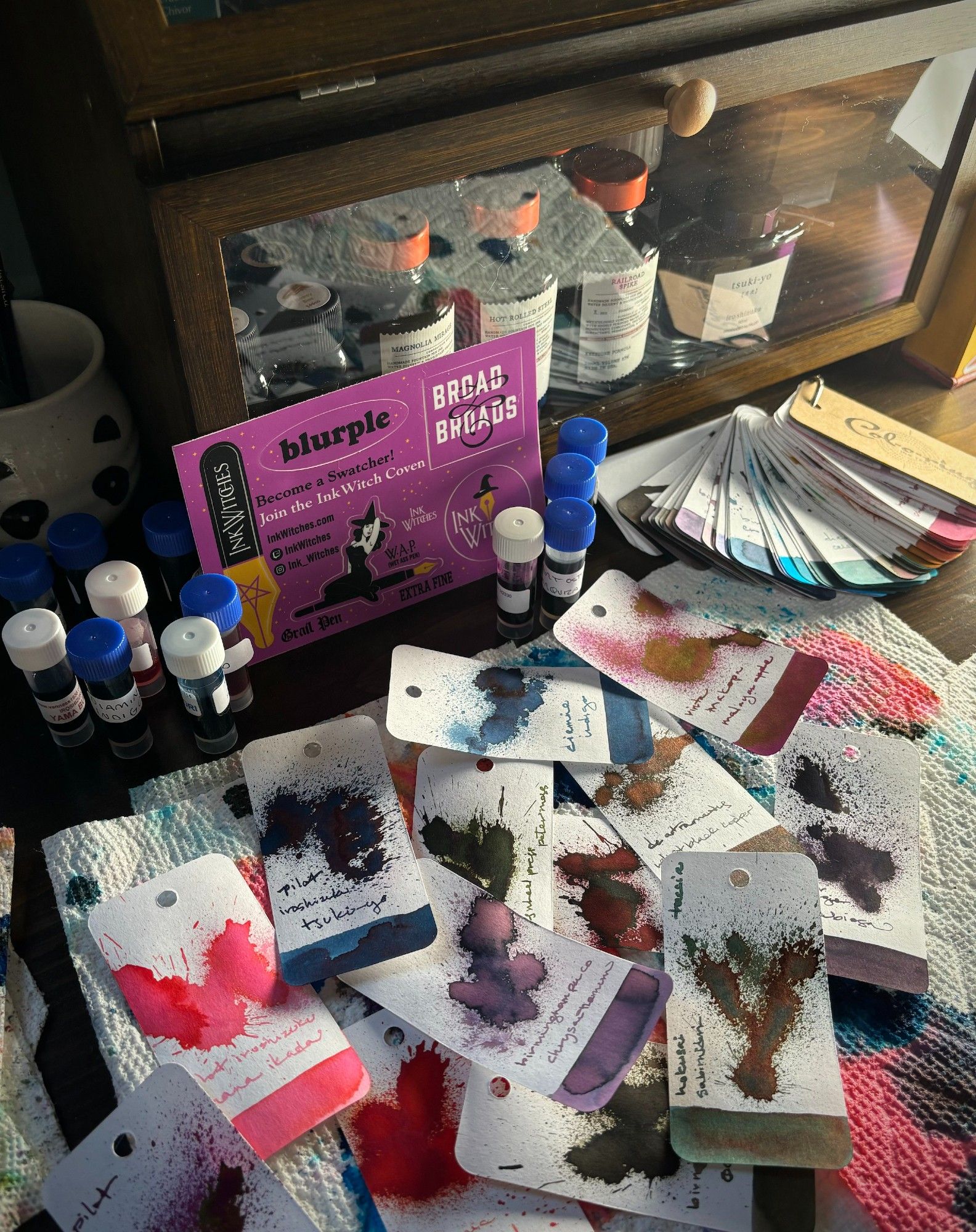 A pile of ink swatch cards sampling fountain pen ink including lots of blues and greens and purples and one bright cherry blossom pink, along with ink sample vials, very messy paper towels and a sheet of purple InkWitches stickers (watch them on twitch!). In the background is a breadbox filled with ink bottles and a cup that looks like a panda.