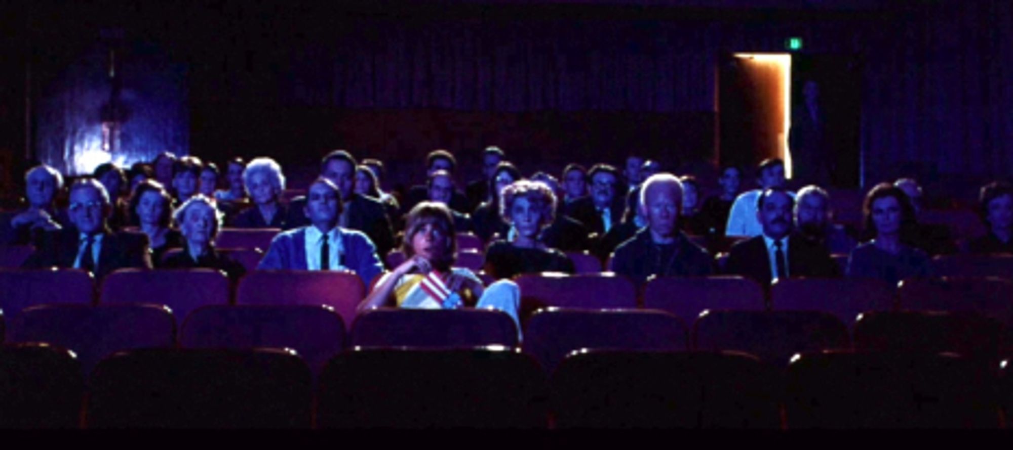 A still from Messiah of Evil. A woman is eating popcorn in the middle of a movie theater. She is surrounded by townsfolk who all look subtly sinister. Right now everyone is keeping up appearances. It's just a normal night at the movies. But soon someone will drop the pretense and things will get nasty quickly.