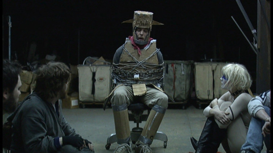 A man in cardboard knight armor is chained to a chair. He's surrounded by several figures sitting on the floor. He's talking to them and they appear to be listening. This is a hostage situation, but it's not going how anyone thought it would.