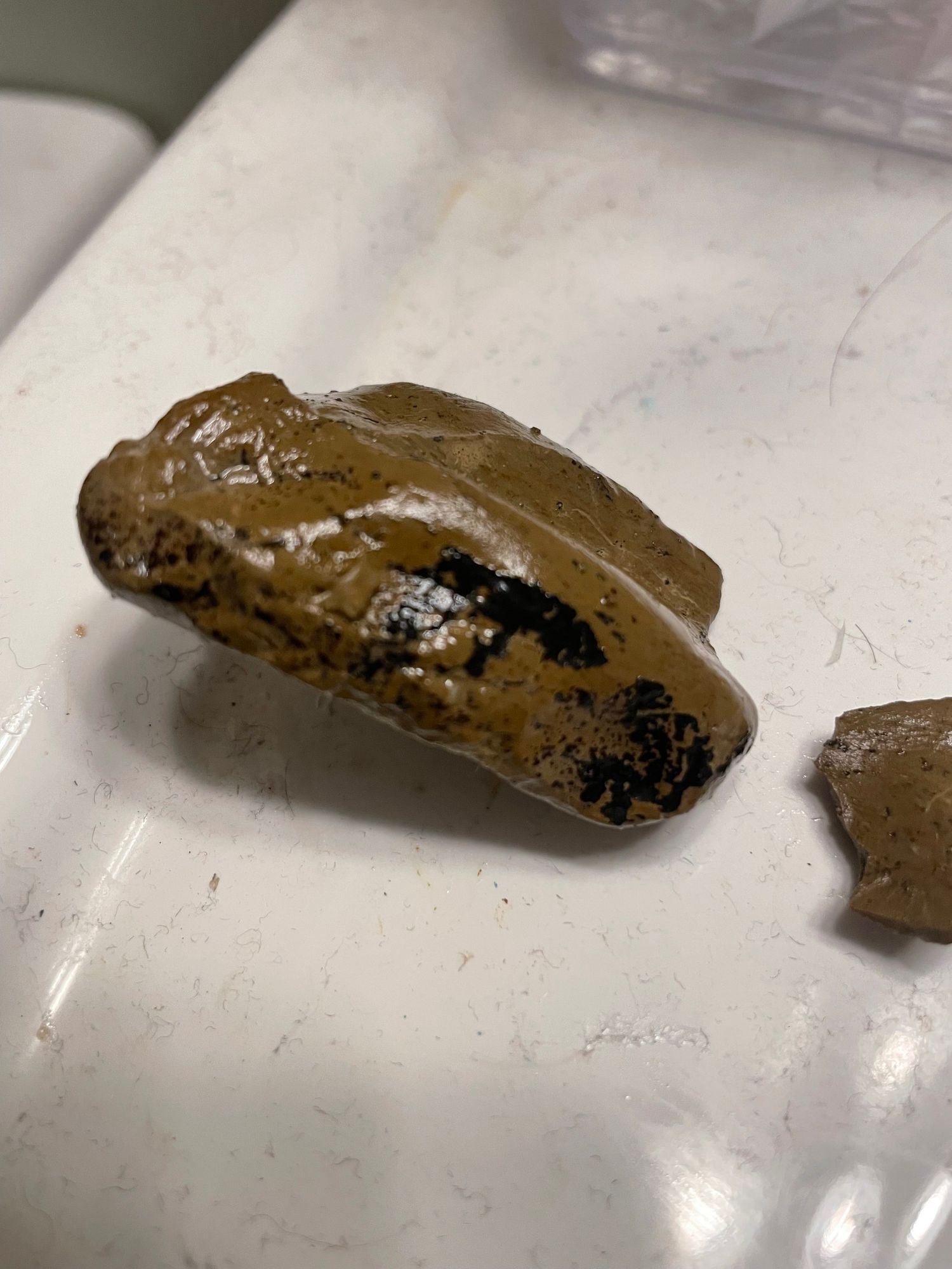 A rock sits on a white sink. It is a sort of rectangular irregular shape. It is mostly brown with black patches. There is a smaller shard to the right.