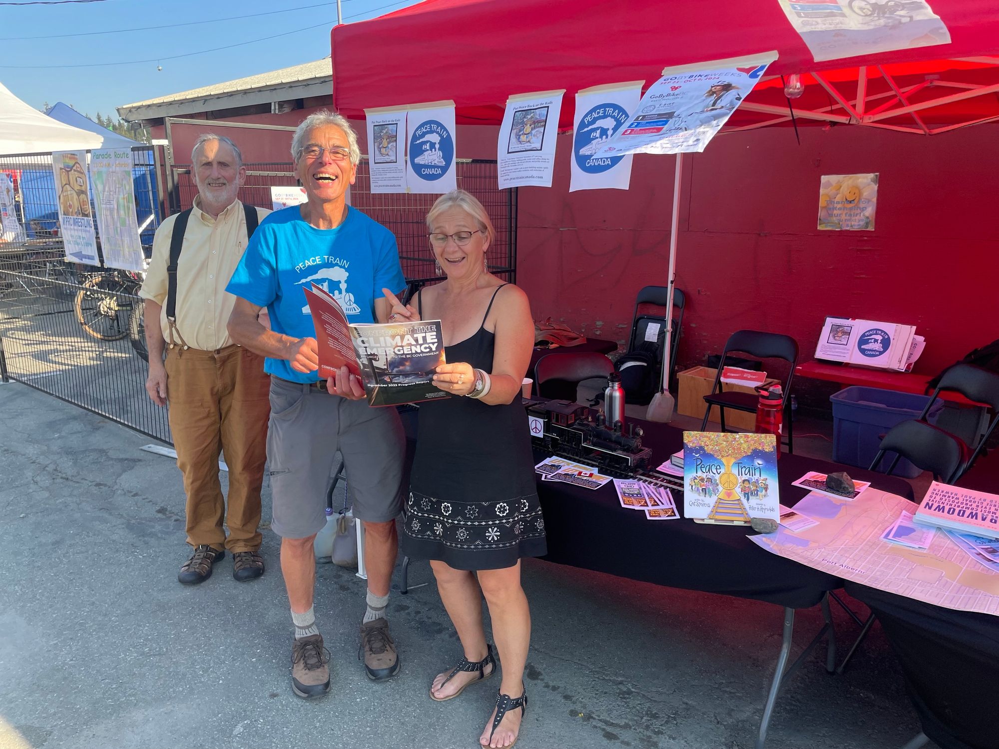 three people pose in front of a booth at the fair. two of them are holding a book about the peace train.