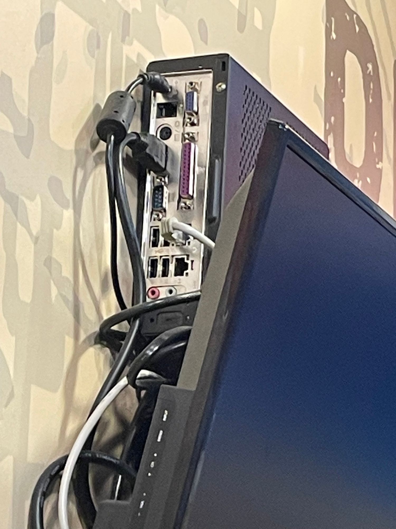 the back of a small pc mounted on a wall behind a screen. There are many ports. 