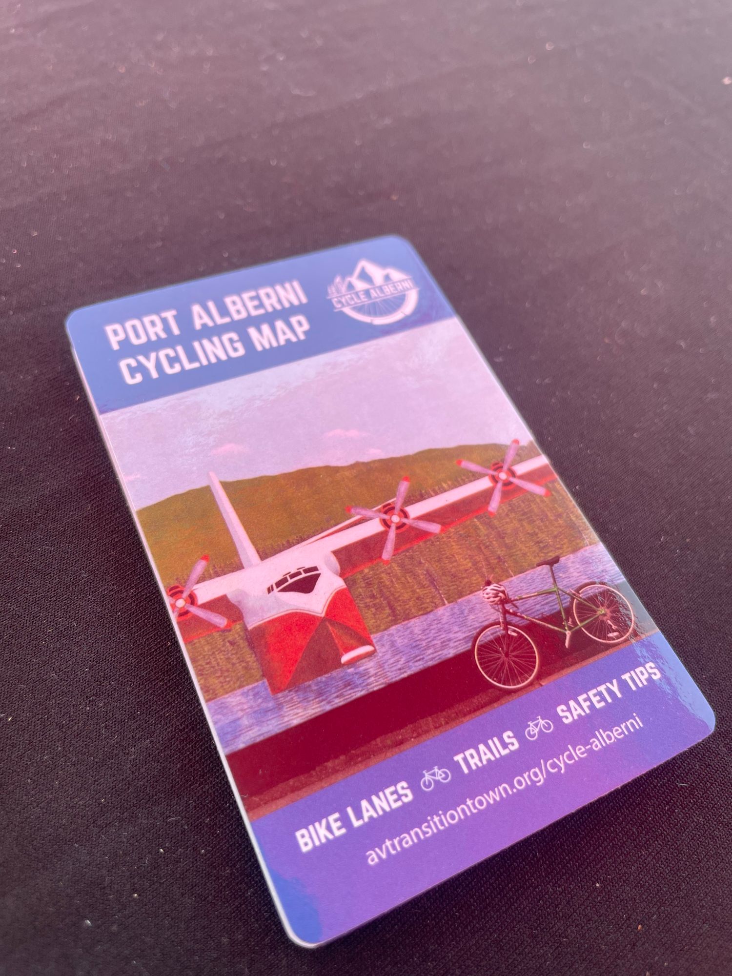 A small rectangular fold out sits on a black table cloth. It says Port Alberni Cycling Map on the bottom it says Bike Lanes Trails and Safety Tips. It has a red and white plane.