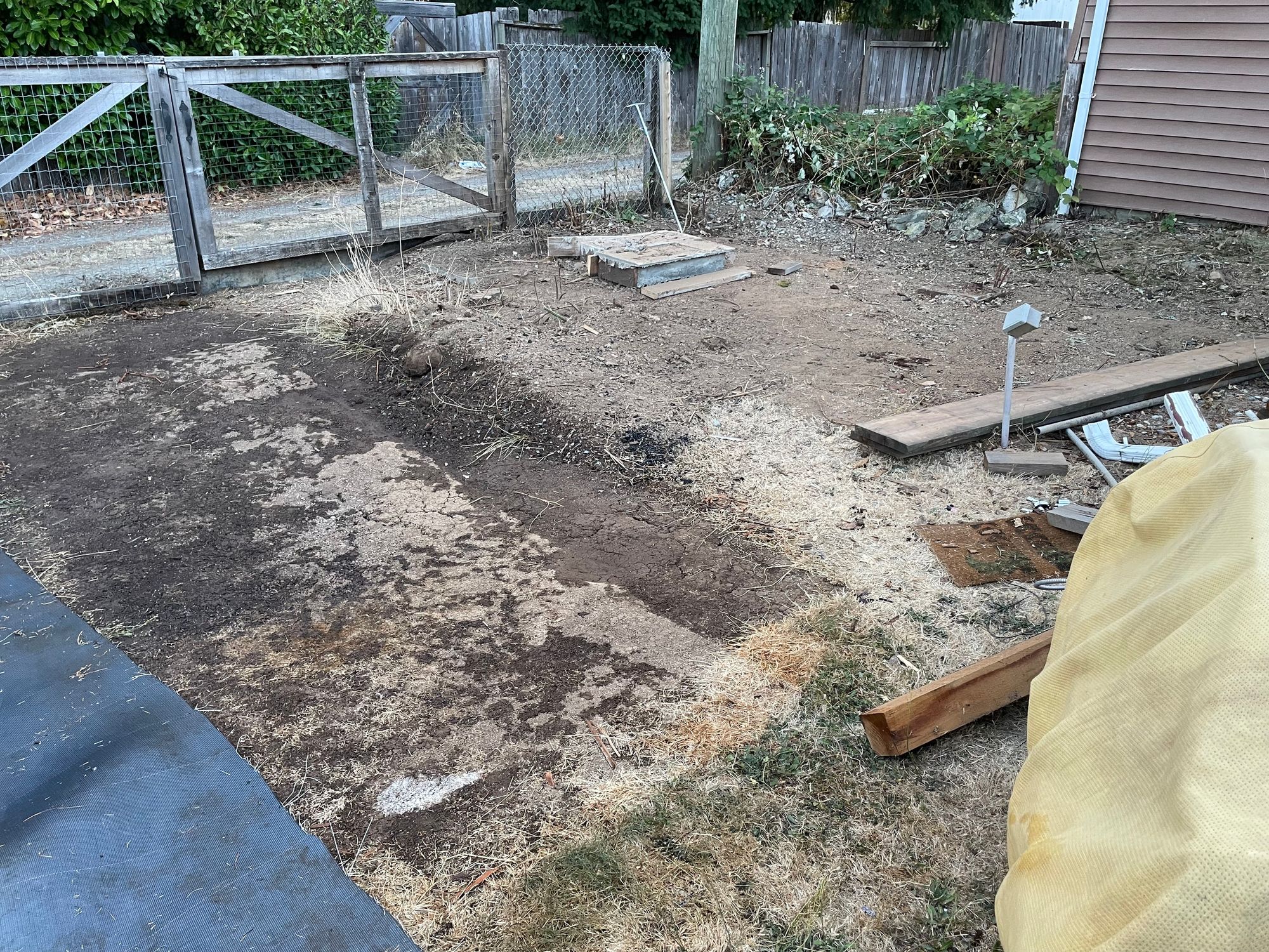 A closer look at the cleared space where the shed was. There is also a section that has been uncovered where the ground is dry and cracked. A blue grey thick fabric like mat is on the ground to the left.