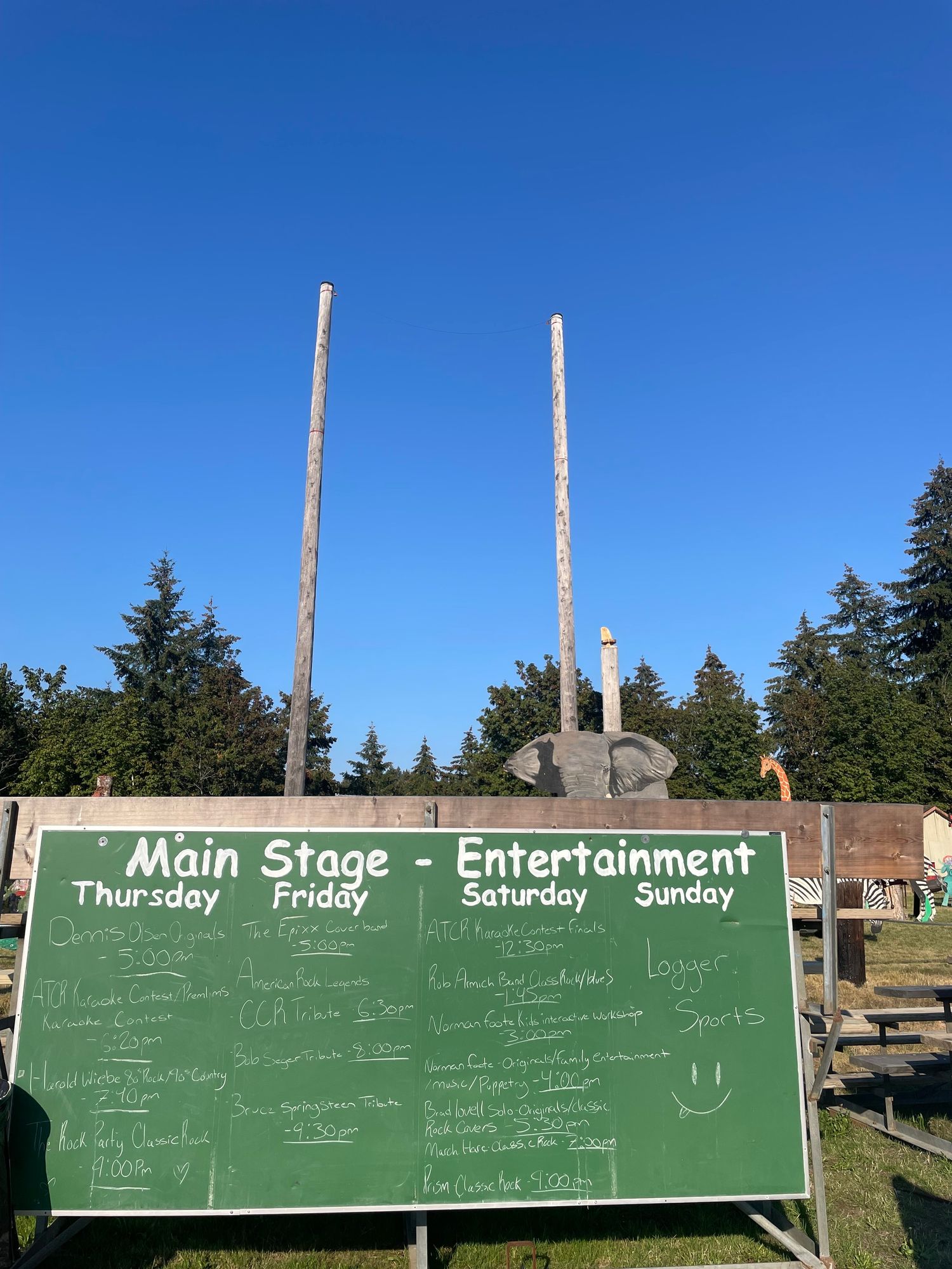 An old green chalkboard has days from Thursday to Sunday with a list of musical acts below them.  
Behind the chalkboard are too very tall, log climbing poles