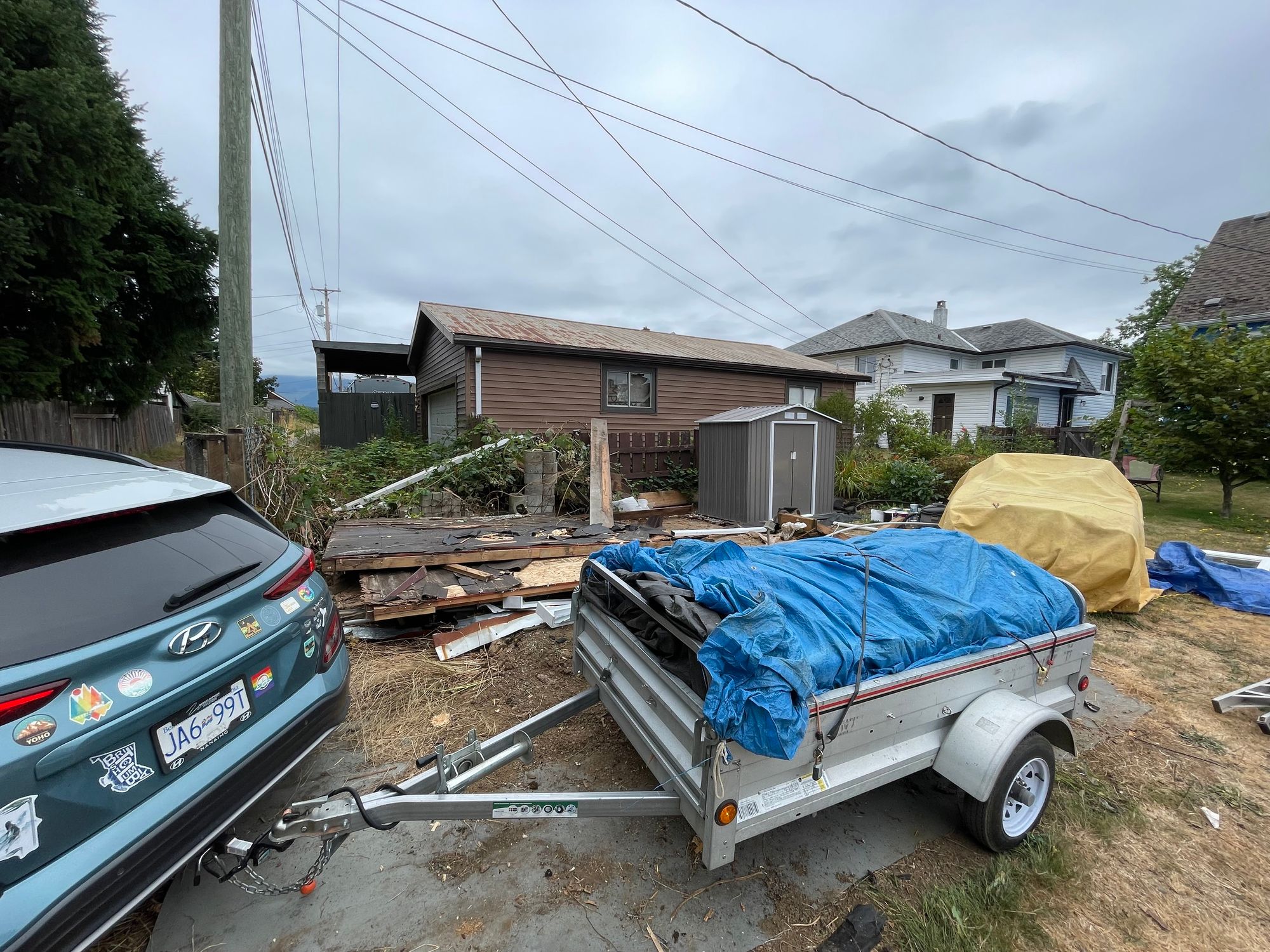 A small car pulling a small trailer that is covered with a blue tarp. Behind is a bunch of debris on the ground from the shed