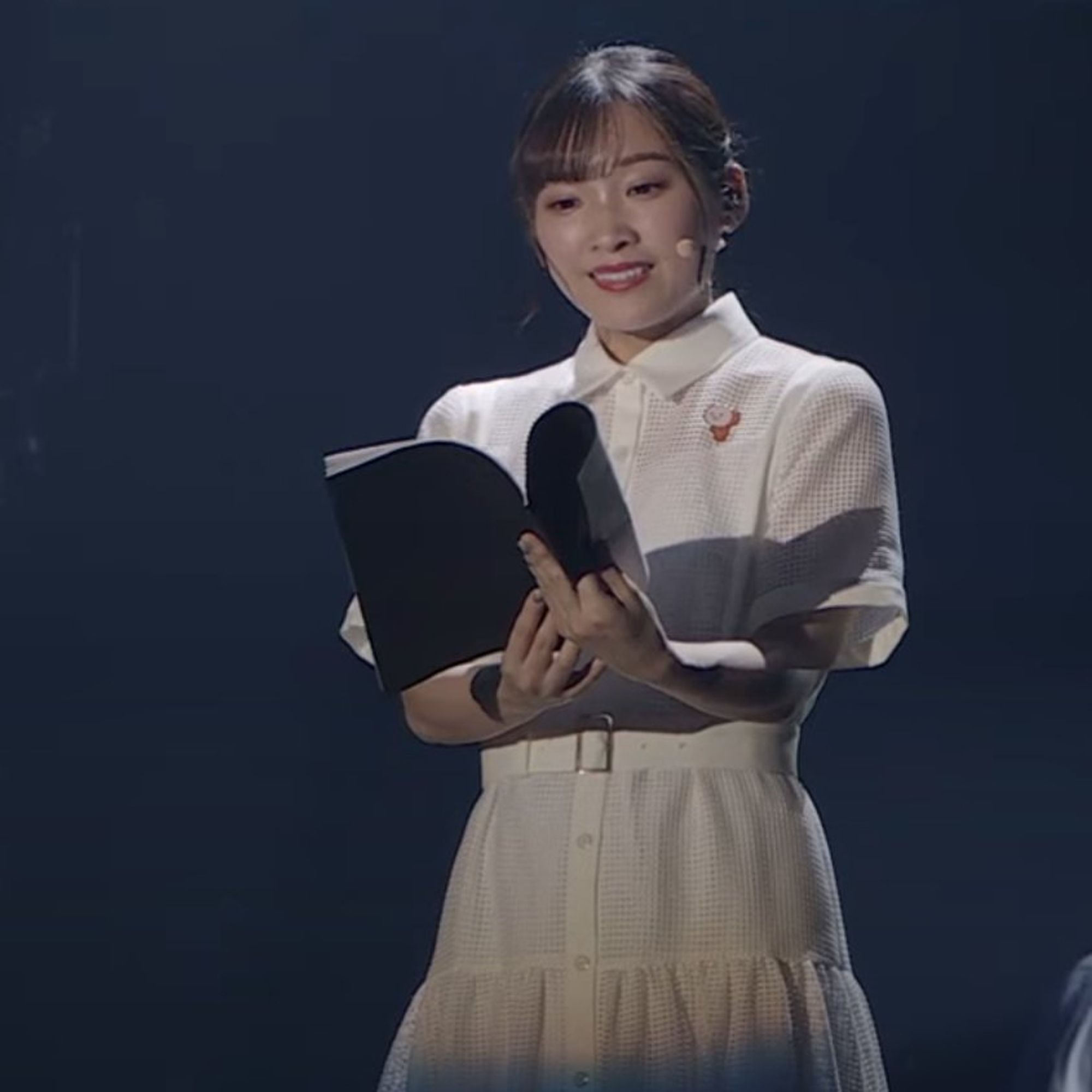 A screenshot from the Mobile Suit Gundam: The Witch From Mercury Festival Asticassia All School Assembly. Kana Ichinose, reading from a black bound script, wears a small pinbadge of the orange hot keychain from G-Witch on her white dress shirt.