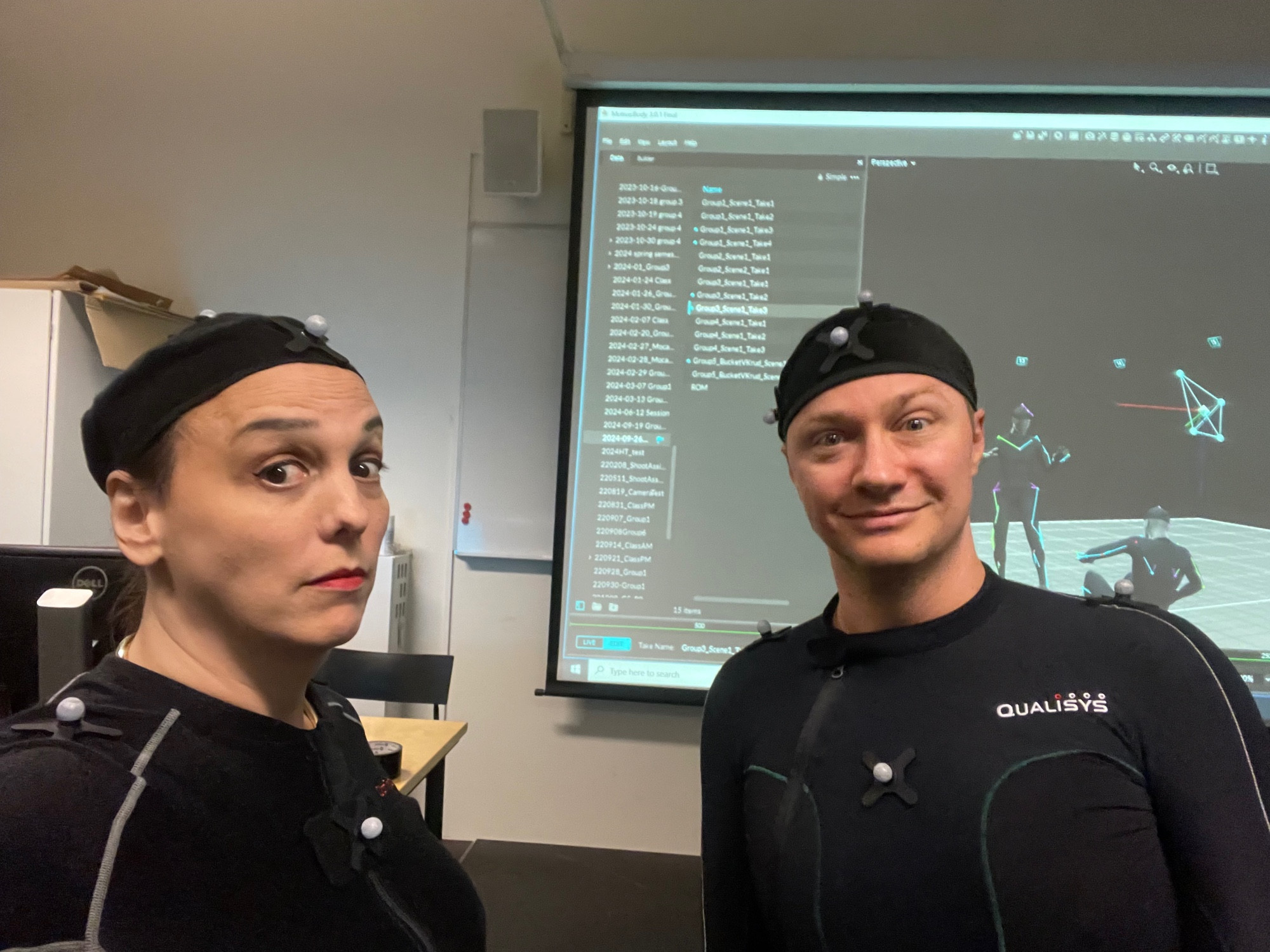 A photograph of two people in motion capture suits in front of a projected computer screen