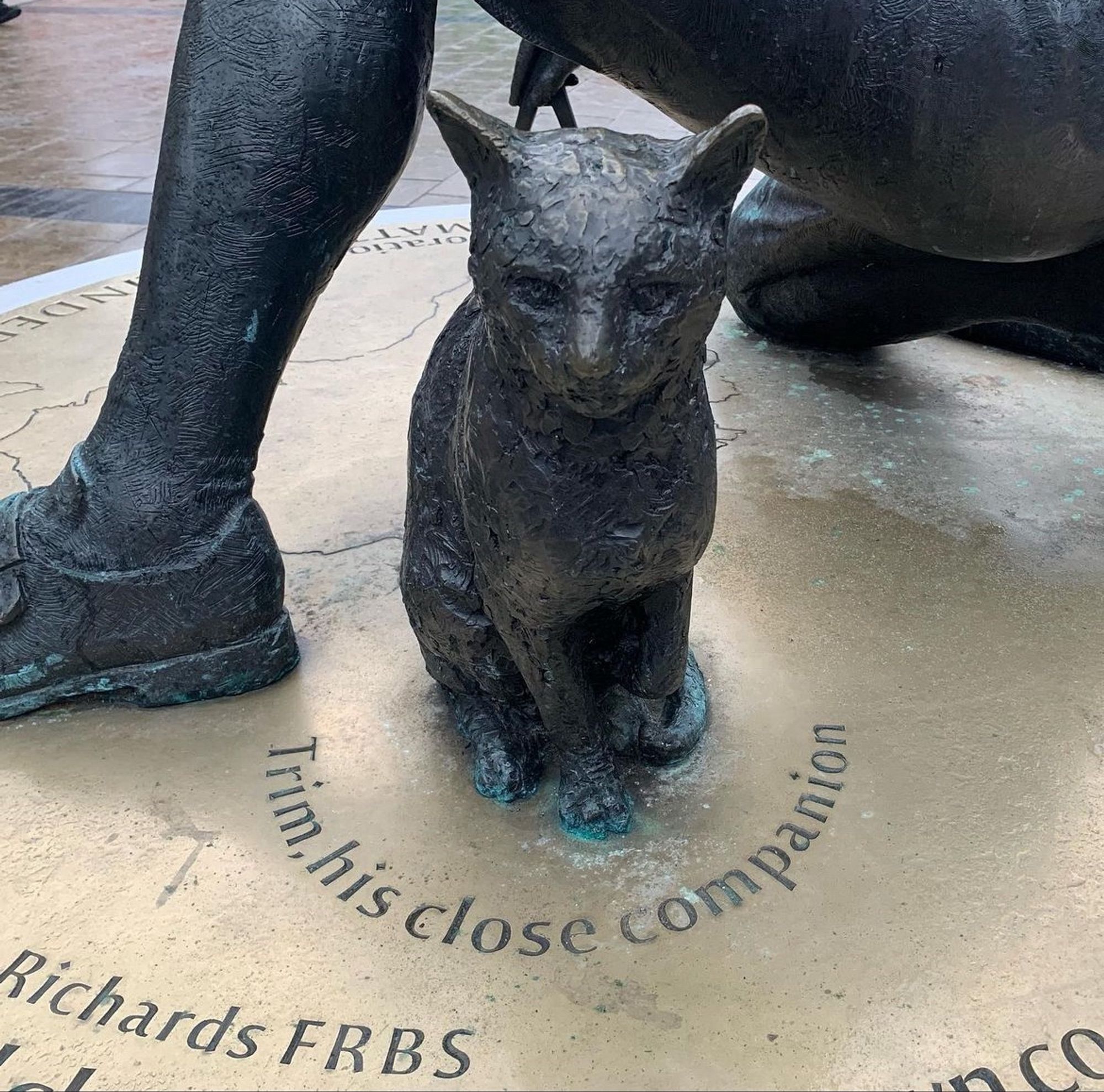 A close-up of the statue of Capt. Matthew Flinders RN at Euston Station. Flinders’s left leg is visible, but the image focuses on a life-size bronze cat, and engraved on the plinth are the words “Trim, his close companion”