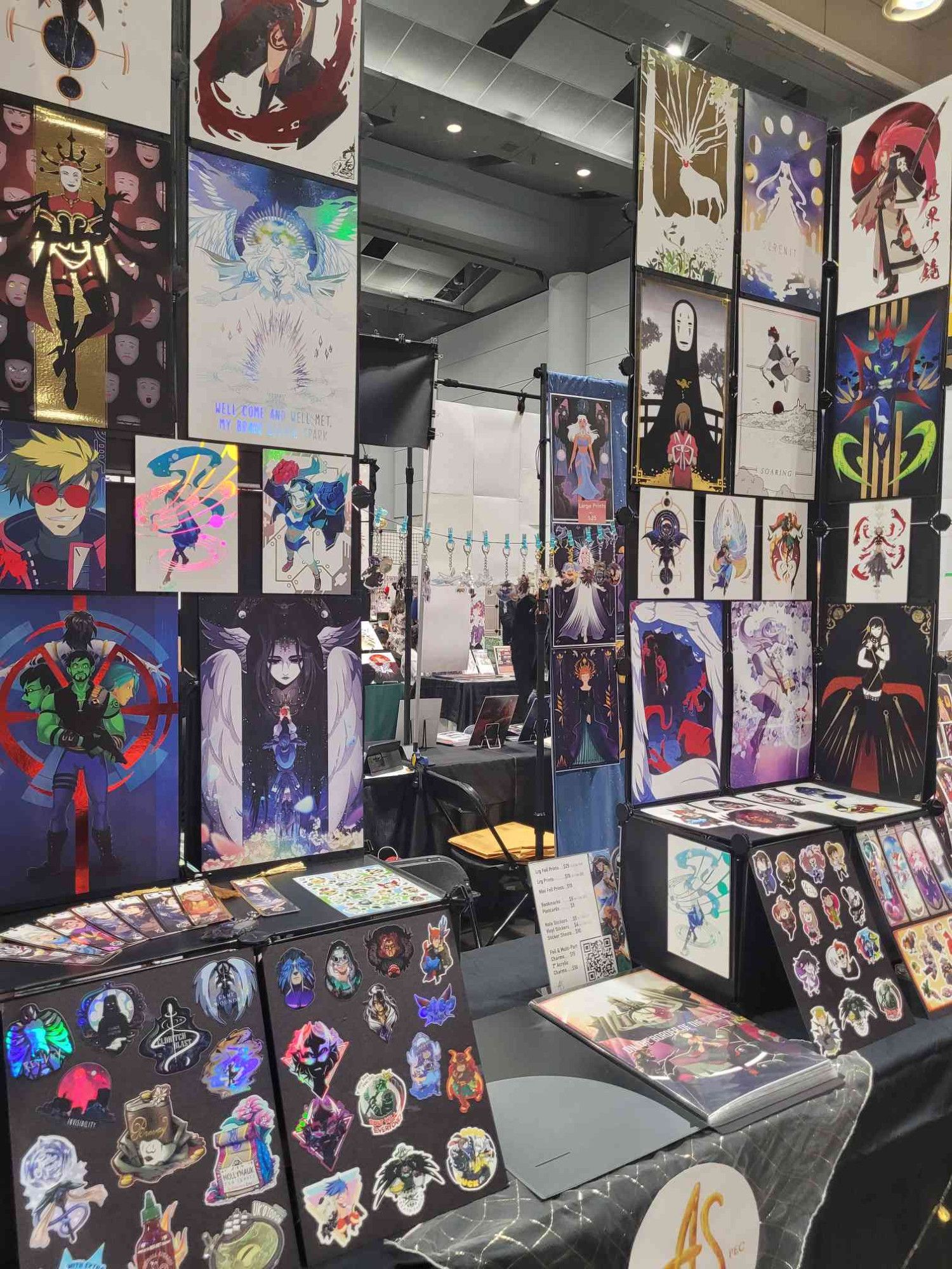 Photo of a convention display in artist alley showcasing our foil prints, stickers, bookmarks and more!!