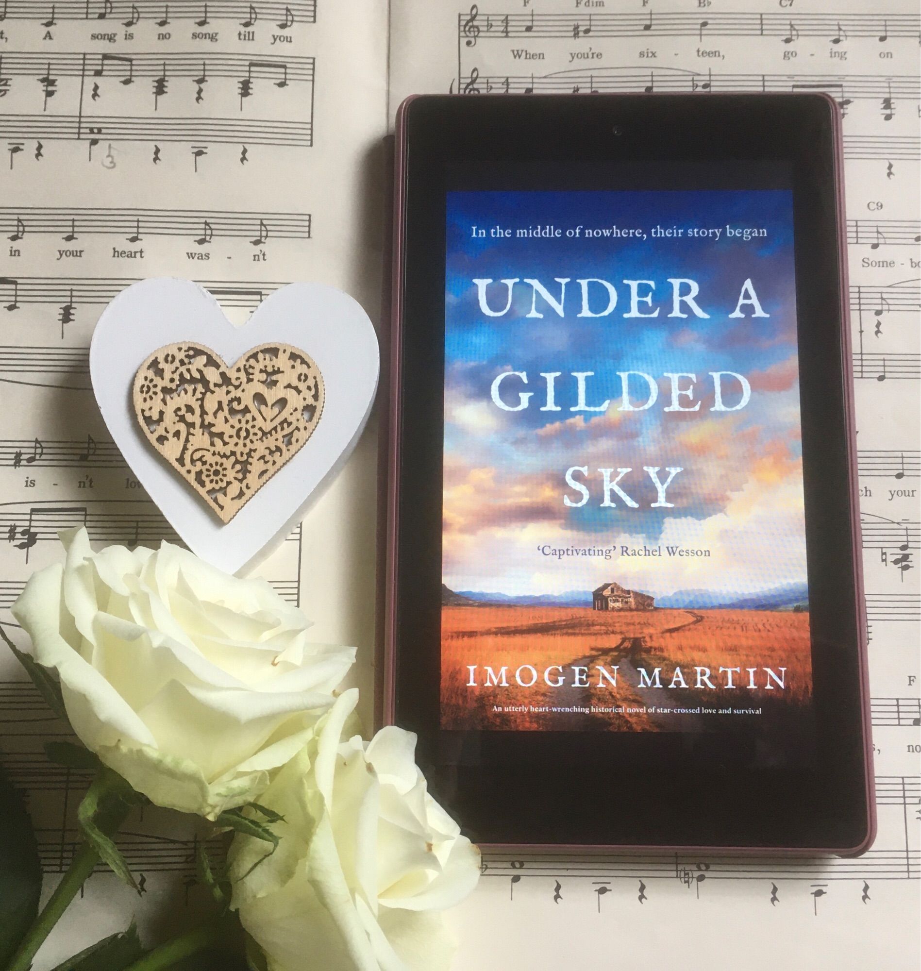 Book cover of Under A Gilded Sky by Imogen Martin on a music score, with heart shape and two white roses