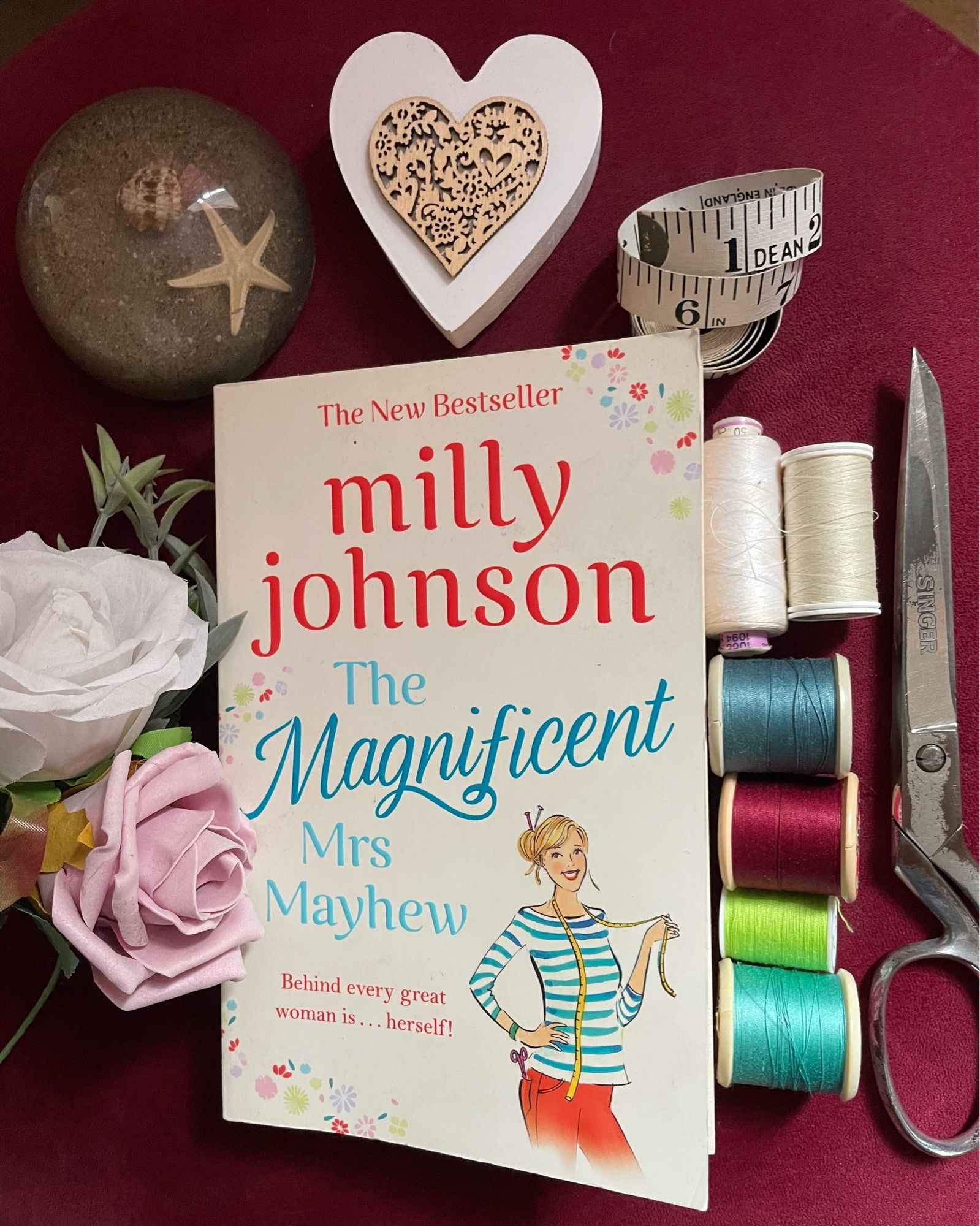 book cover of The Magnificent Mrs Mayhew by Milly Johnson surrounded by cotton reels, tape measure, scissors, heart shape shell and sand paperweight, and two flowers