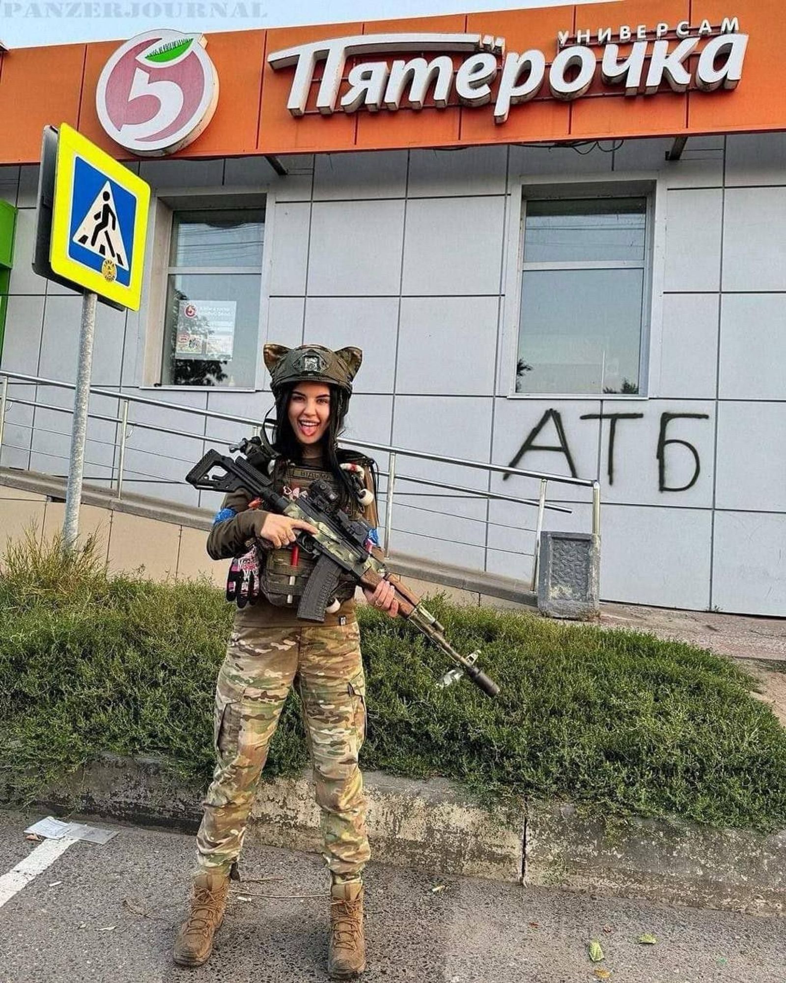 A woman in military camouflage stands in front of a "Pyaterochka" supermarket, a well-known Russian grocery store chain, as indicated by the signage above her. She is smiling and holding a rifle, dressed in full tactical gear, including a helmet with cat ears attached to it. The helmet has various patches and accessories, including what appear to be magazines and a radio. A pedestrian crossing sign is visible to her left, and behind her, the wall of the store has the graffiti "ATБ" spray-painted in black. The overall scene combines elements of military imagery with a civilian setting.