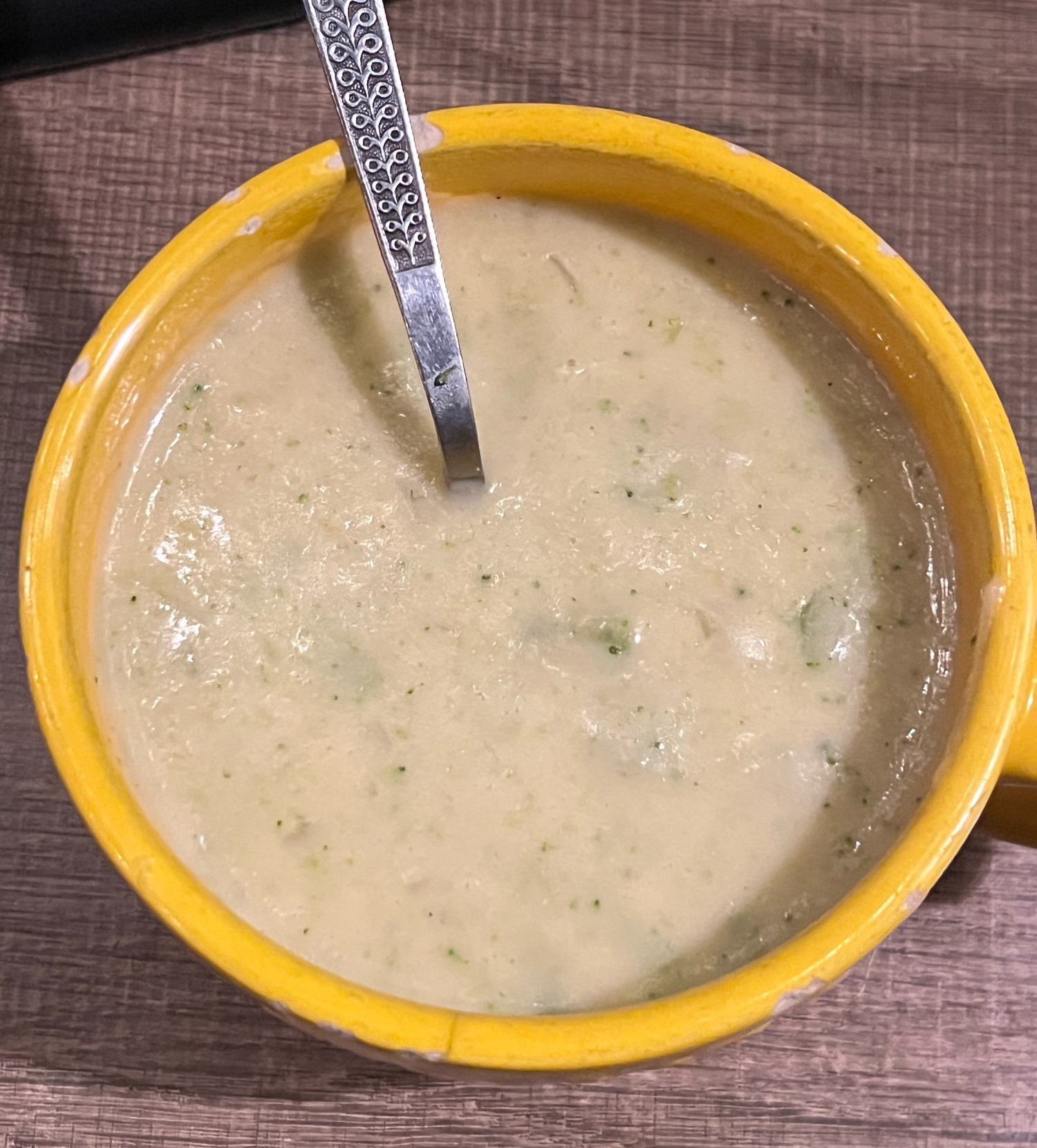 A creamy yellow broccoli cheddar soup in a yellow soup mug that has seen better days and has a few chips in the rim