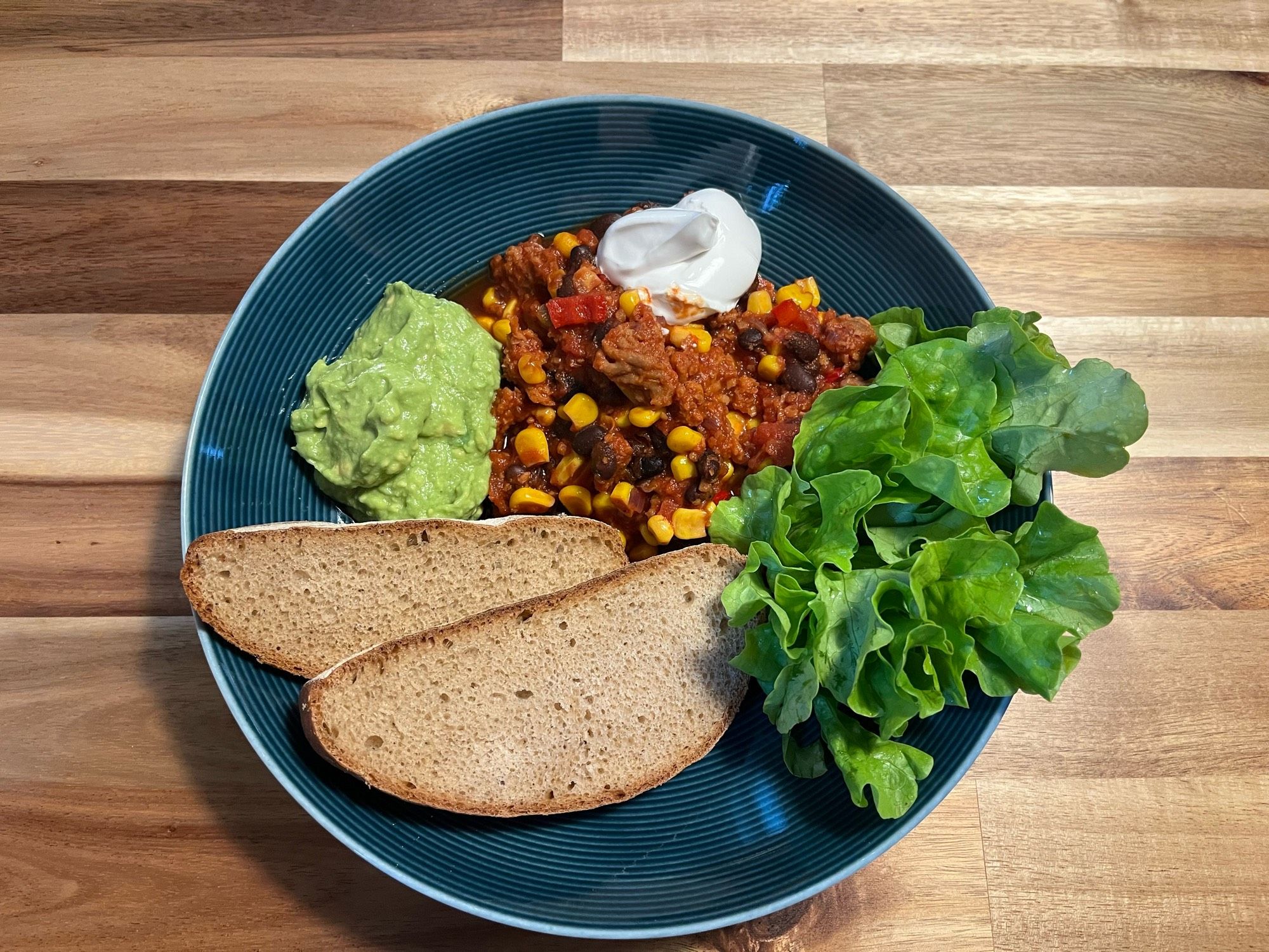 Ein blauer Teller mit veganem Chili sin Carne, einem Klecks veganem Rahm und Guacamole, zwei Scheiben Brot und ein paar Salatblättern.