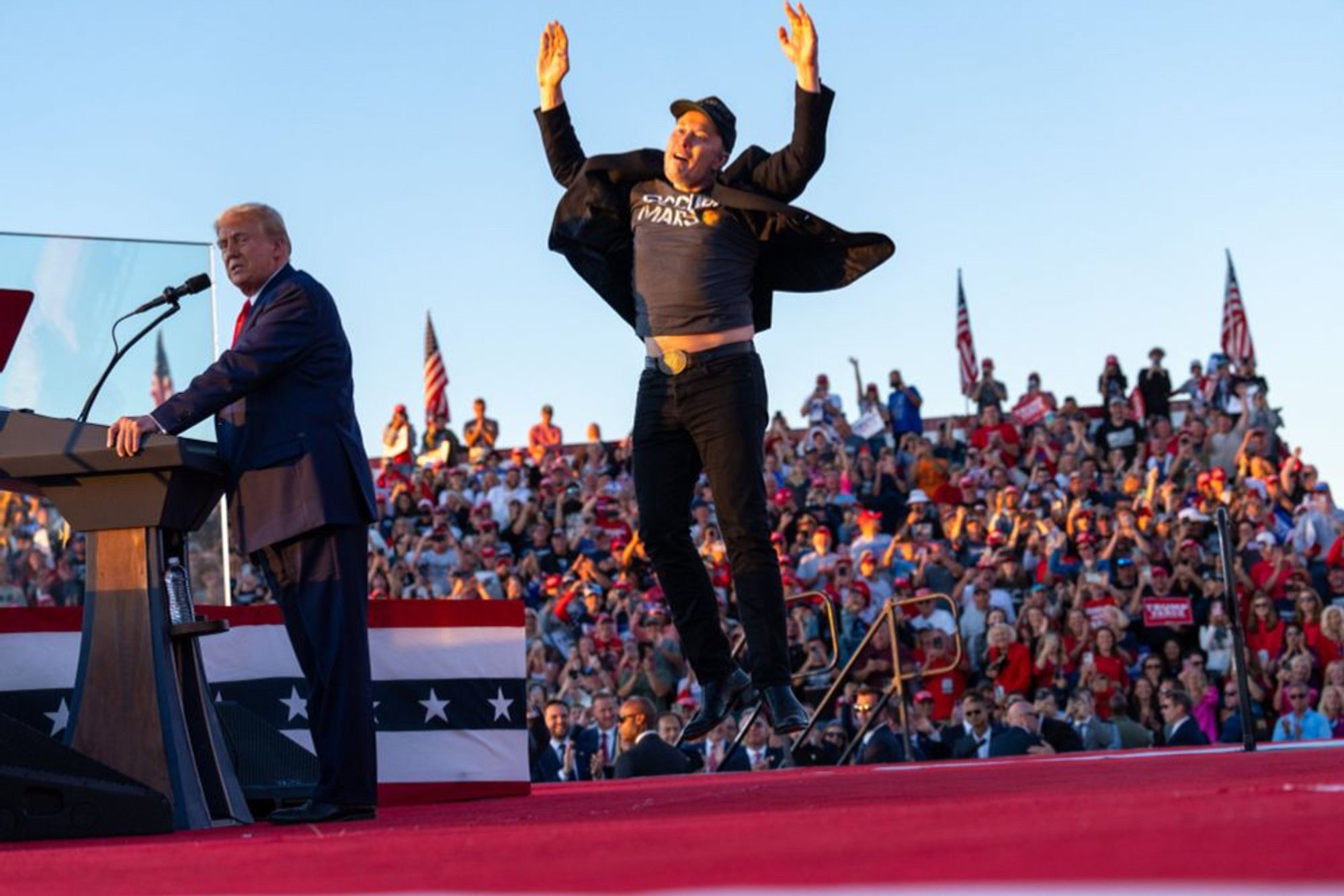 Elon Musk jumping in the air with hands raised in celebration behind Donald Trump at a MAGA rally.