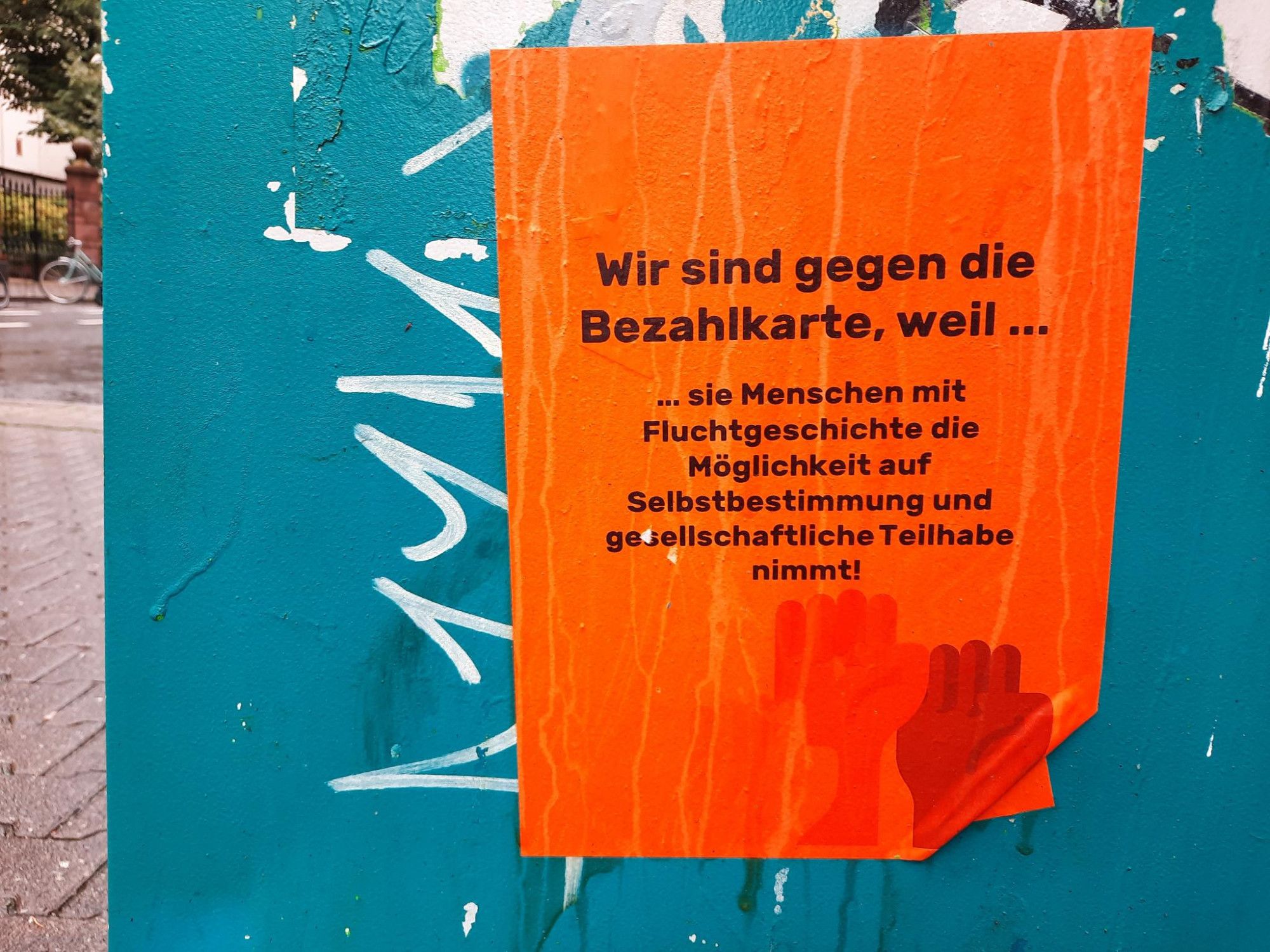 Auf orangem Textfeld in schwarzer Schrift: „Wir sind gegen die Bezahlkarte, weil sie Menschen mit Fluchtgeschichte die Möglichkeit auf Selbstbestimmung und gesellschaftliche Teilhabe nimmt!“. Darunter drei in die Höhe gestreckte Fäuste in den Farben Altrosa, Sandbraun und Dunkelbraun. Angebracht ist das Plakat an einem anderen, blau bemalten Stromkasten. Alles ist naß, es regnet.