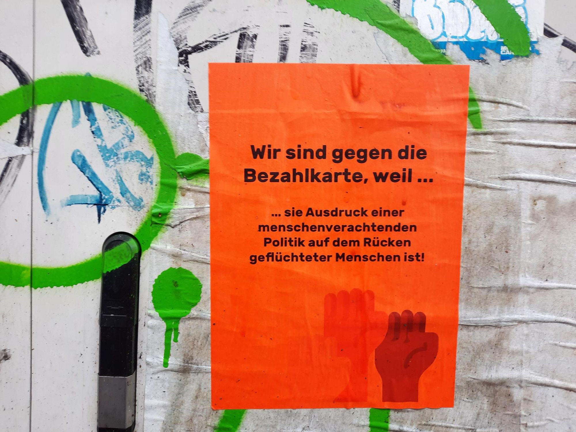 Auf orangem Textfeld in schwarzer Schrift: „Wir sind gegen die Bezahlkarte, weil sie Ausdruck einer menschenverachtenden Politik auf dem Rücken geflüchteter Menschen ist!“. Darunter drei in die Höhe gestreckte Fäuste in den Farben Altrosa, Sandbraun und Dunkelbraun. Angebracht ist das Plakat an einem Stromkasten auf die Kreise und Halbkreise mit grüner Farbe gemalt sind.  Alles ist naß, es regnet.