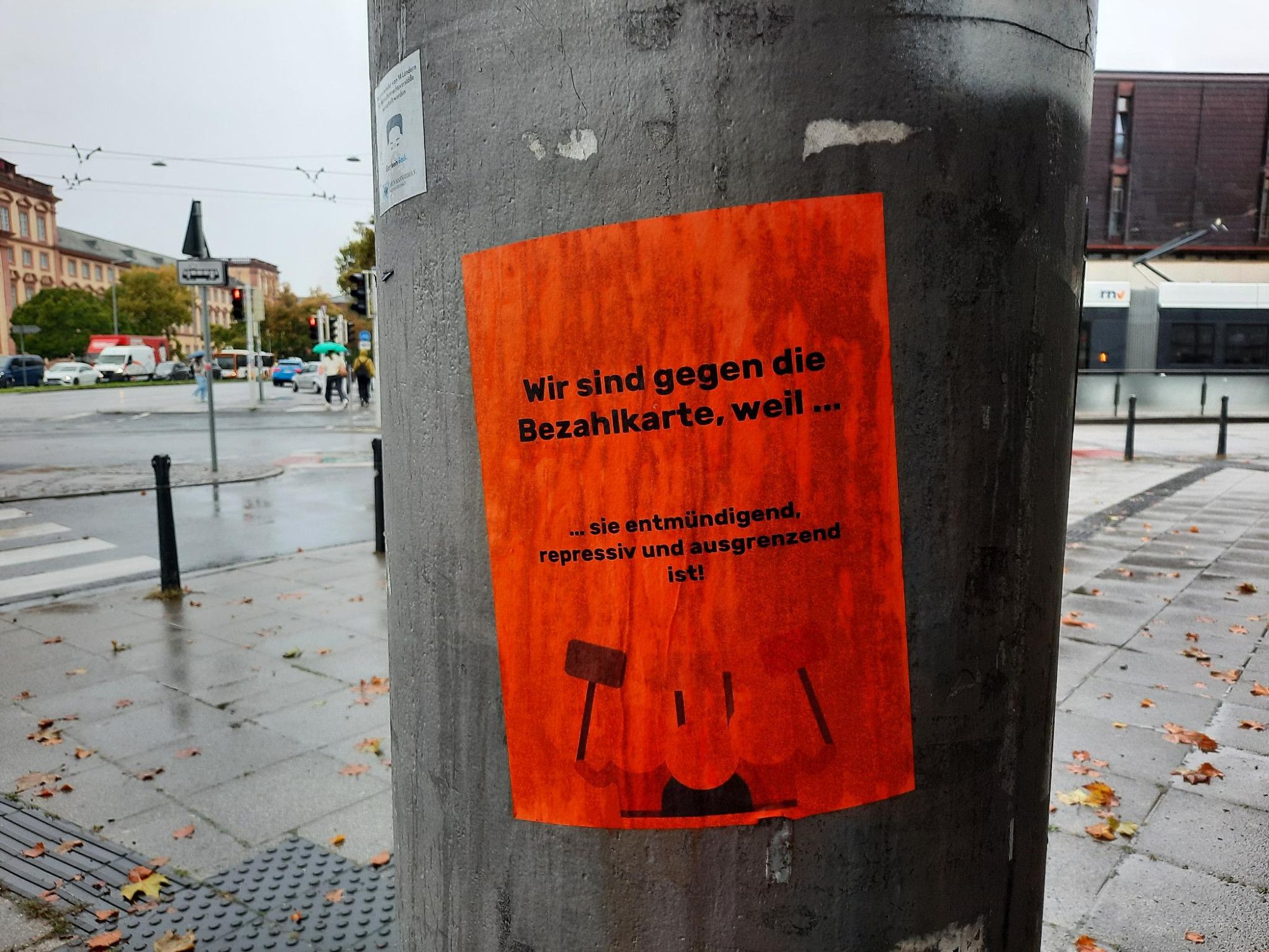 Auf orangem Textfeld in schwarzer Schrift: „Wir sind gegen die Bezahlkarte, weil sie entmündigend, repressiv und ausgrenzend ist!“. Darunter drei Piktogramm-Menschen, die vier Schilder in die Höhe halten. Die Gesichter der Piktogramm- Menschen sind Mendelweiß und in heller Lachsfarbe, ihre Kleidung ist hell -bis dunkelbraun. Die Schilder, alle auf dunkelblauen Stäben, sind Kornblumenblau, aus blassem Türkis, helle Senffarbe und Rosa.
Das Plakat ist an einem Laternenpfahl angebracht. Links im Hintergrund Teile vom Schloss, Autos, Menschen mit Regenschirm und rechts vom Laternenpfahl Teile vom Gericht. Plakat, Straße, alles naß, denn es regnet.