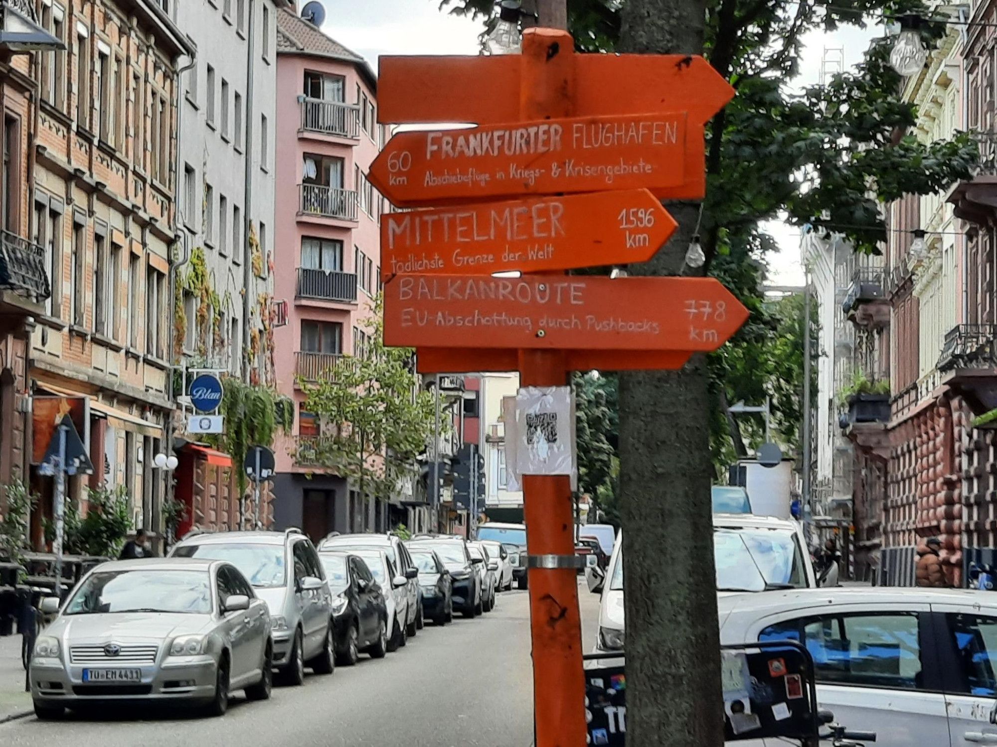 An orangem Stamm befinden sich orange Pfeile, die mit weißer Schrift folgende Orte samt Kilometerangabe hinweisen: Frankfurt Flughafen, Abschiebeflüge in Kriegs – und Krisengebiete, 60 km. Mittelmeer, tödlichste Grenze der Welt, 1596 km. Balkanroute, EU - Abschottung durch Pushbacks (gewaltvolles, oft tödliches zurückstoßen an den EU-Grenzen), 778 km (mehr Pfeile sind nicht zu erkennen). Unterhalb der Pfeile befindet sich ein QR-Code mit weiteren Infos. Im Hintergrund Bäume, rechts Autos auf Parkplätzen auf dem Bürgersteig und links auf der Straße. Häuser, vereinzelt Schilder an Häusern die auf Kneipen und Imbisse hinweisen.