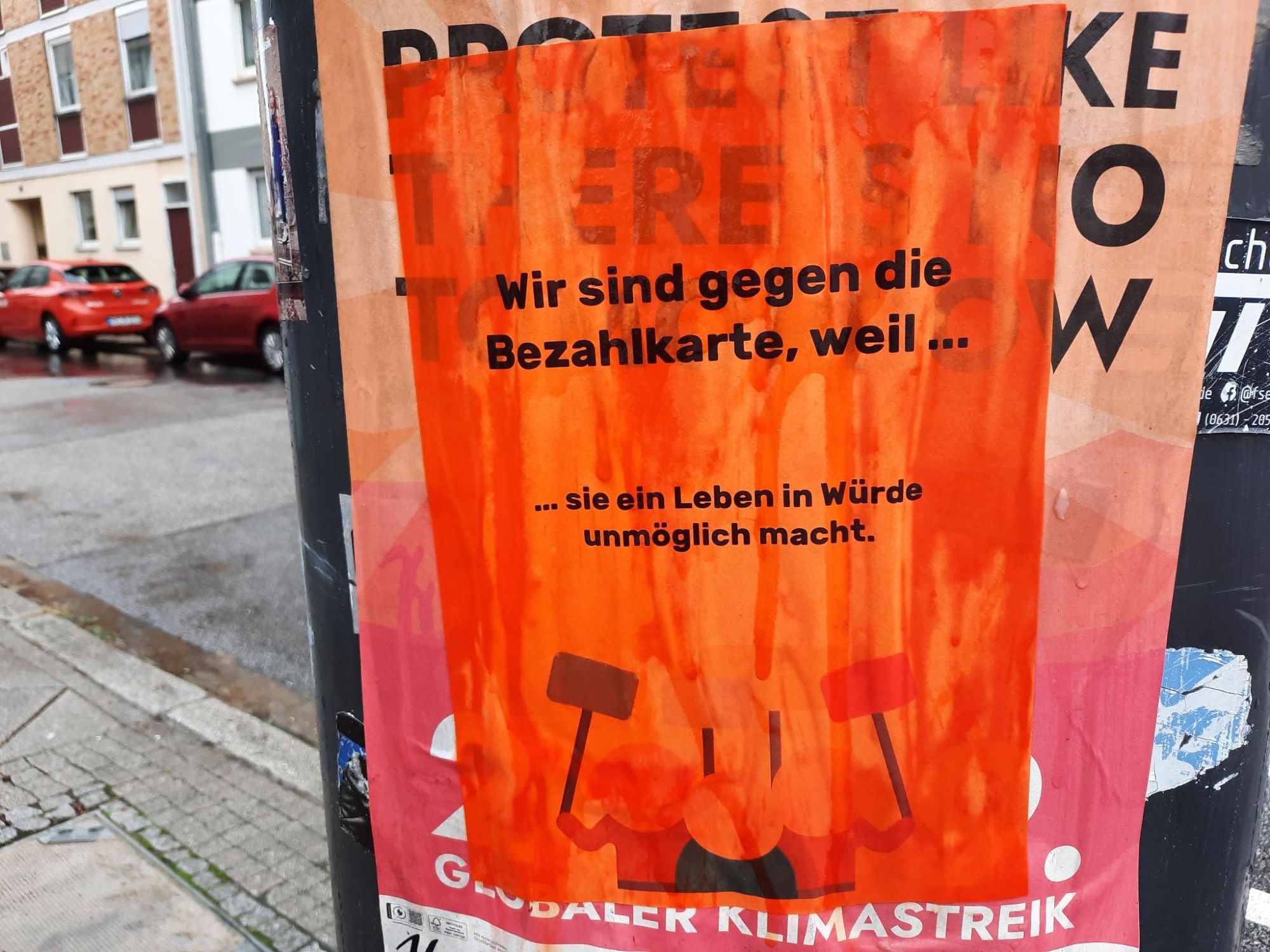 Auf orangem Textfeld in schwarzer Schrift: „Wir sind gegen die Bezahlkarte, weil sie ein Leben in Würde unmöglich macht!“. Darunter drei Piktogramm-Menschen, die vier Schilder in die Höhe halten. Die Gesichter der Piktogramm- Menschen sind Mendelweiß und in heller Lachsfarbe, ihre Kleidung ist hell -bis dunkelbraun. Die Schilder, alle auf dunkelblauen Stäben, sind Kornblumenblau, aus blassem Türkis, helle Senffarbe und Rosa. Das Plakat ist angemacht an einem Papierkorb. Im Hintergrund Autos und Häuser. Alles naß, es regnet.