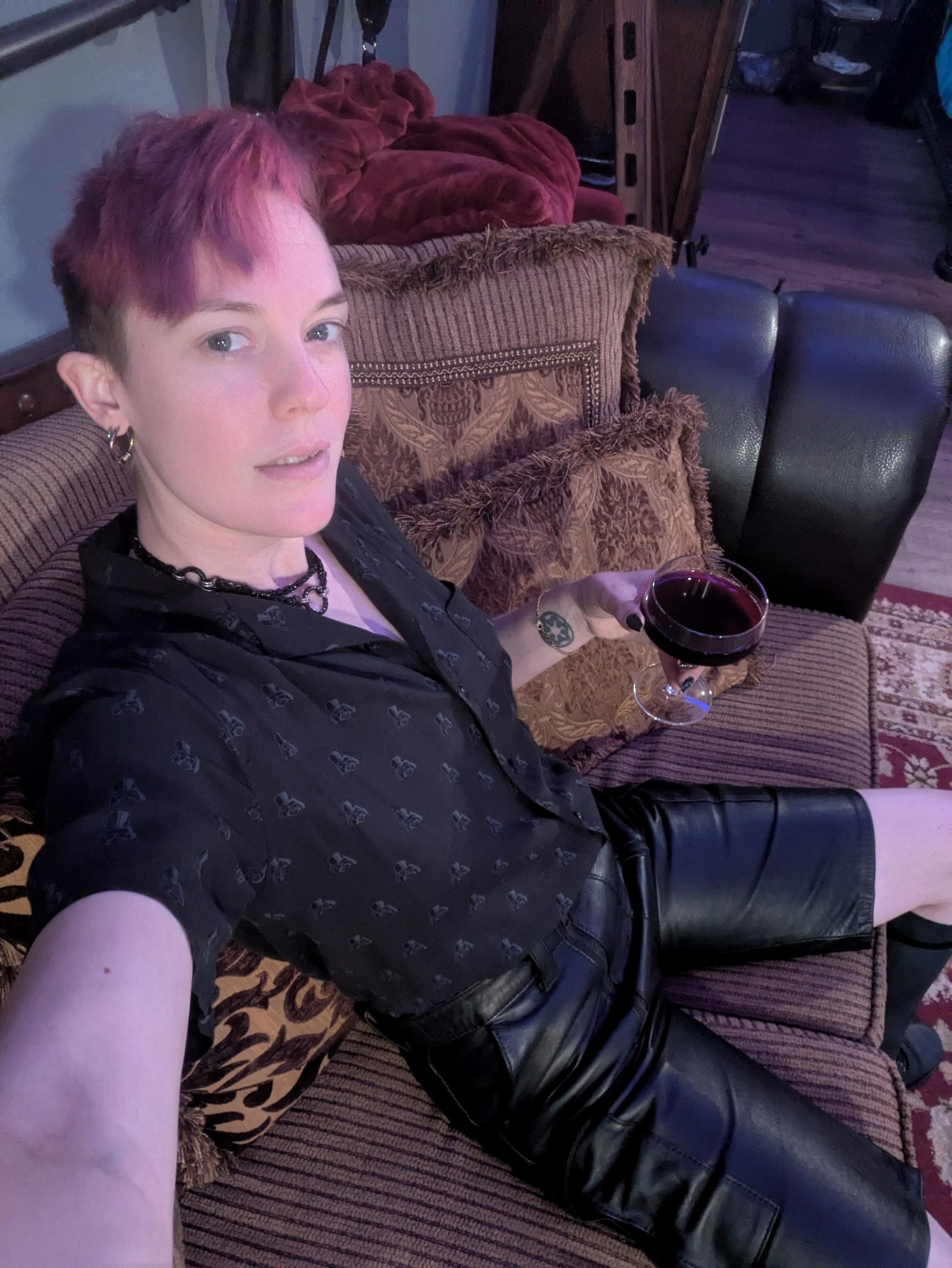 Delia reclines on a couch wearing black leather cargo shorts, a short sleeved button down with Darth Vader step and repeat pattern, and a coupe glass in hand